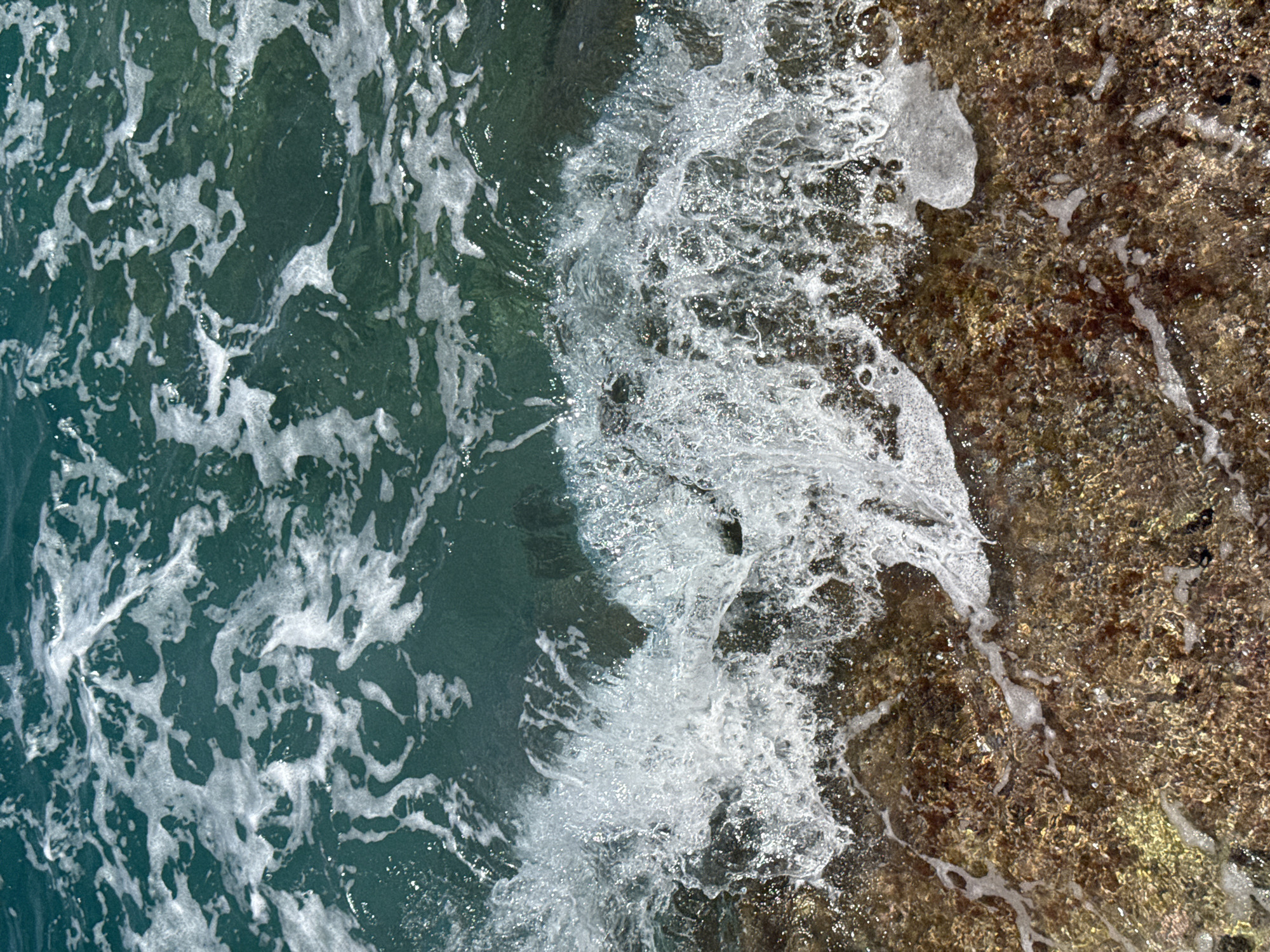 The image captures a close-up, overhead view of shallow, clear turquoise water lapping against a rocky shoreline. The water exhibits foamy white crests and turbulent movement as it interacts with the submerged rocks, which are textured and reddish-brown. Sunlight glints off the water's surface, creating bright highlights and shimmering effects. The scene is devoid of any human presence or man-made structures, suggesting a natural coastal environment. The location is identified as Dubraljevina, Montenegro.