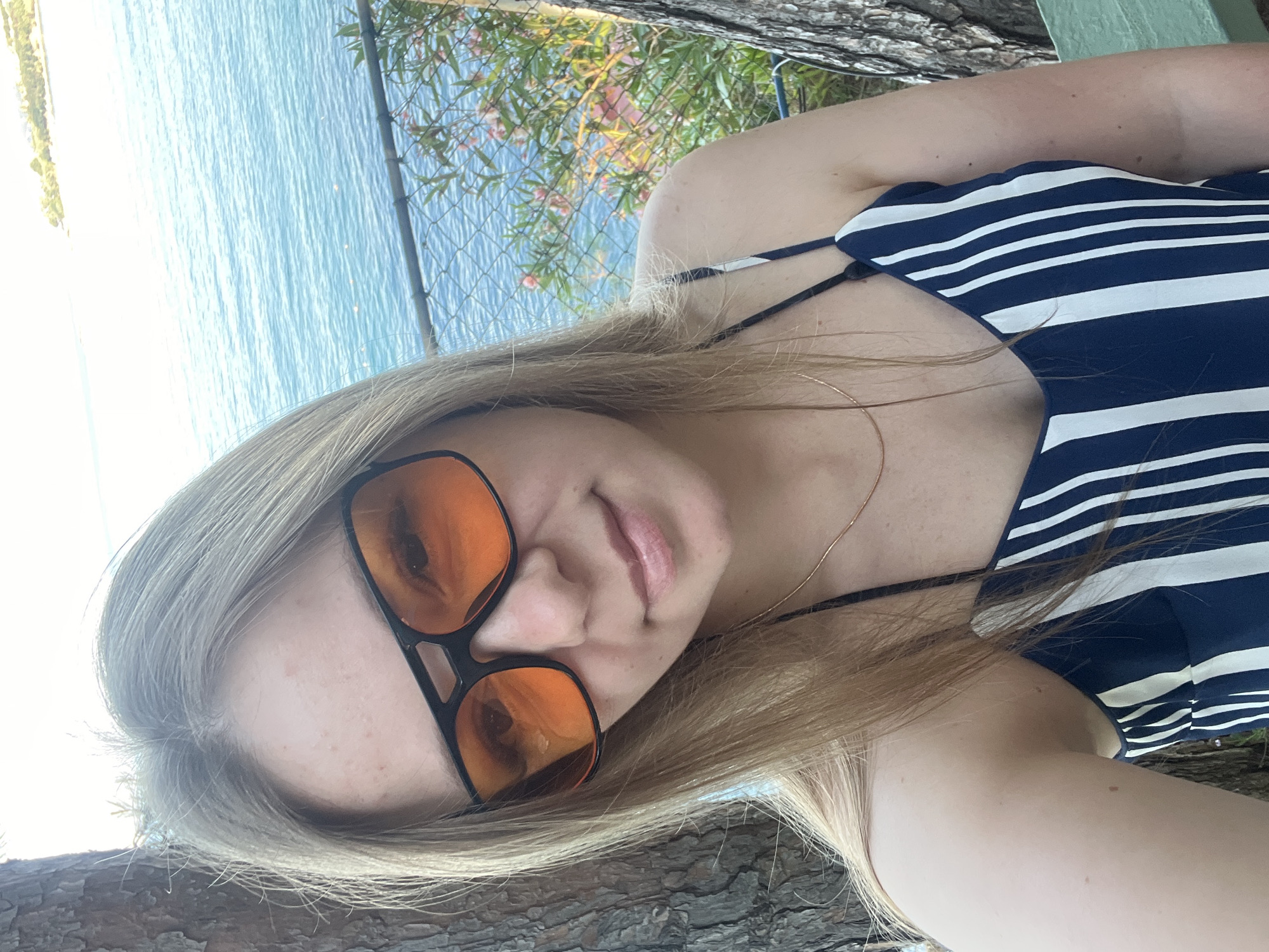 A young woman with blonde hair and orange-tinted sunglasses is pictured in a close-up selfie. She is wearing a navy and white striped dress and a thin gold necklace. Her hair is shoulder-length and styled down. Behind her, a body of blue water, possibly the Mediterranean Sea, is visible. In the background, there is also a blurred island or coastline with some vegetation and buildings. The scene appears to be outdoors, likely in Alanya, Turkey, given the coastal setting and vegetation typical of the region. The lighting suggests it is daytime, possibly late afternoon, with the sun casting a bright light. There is a rustic tree trunk to the left of the frame and another to the right. A green bench is partially visible in the lower right corner.