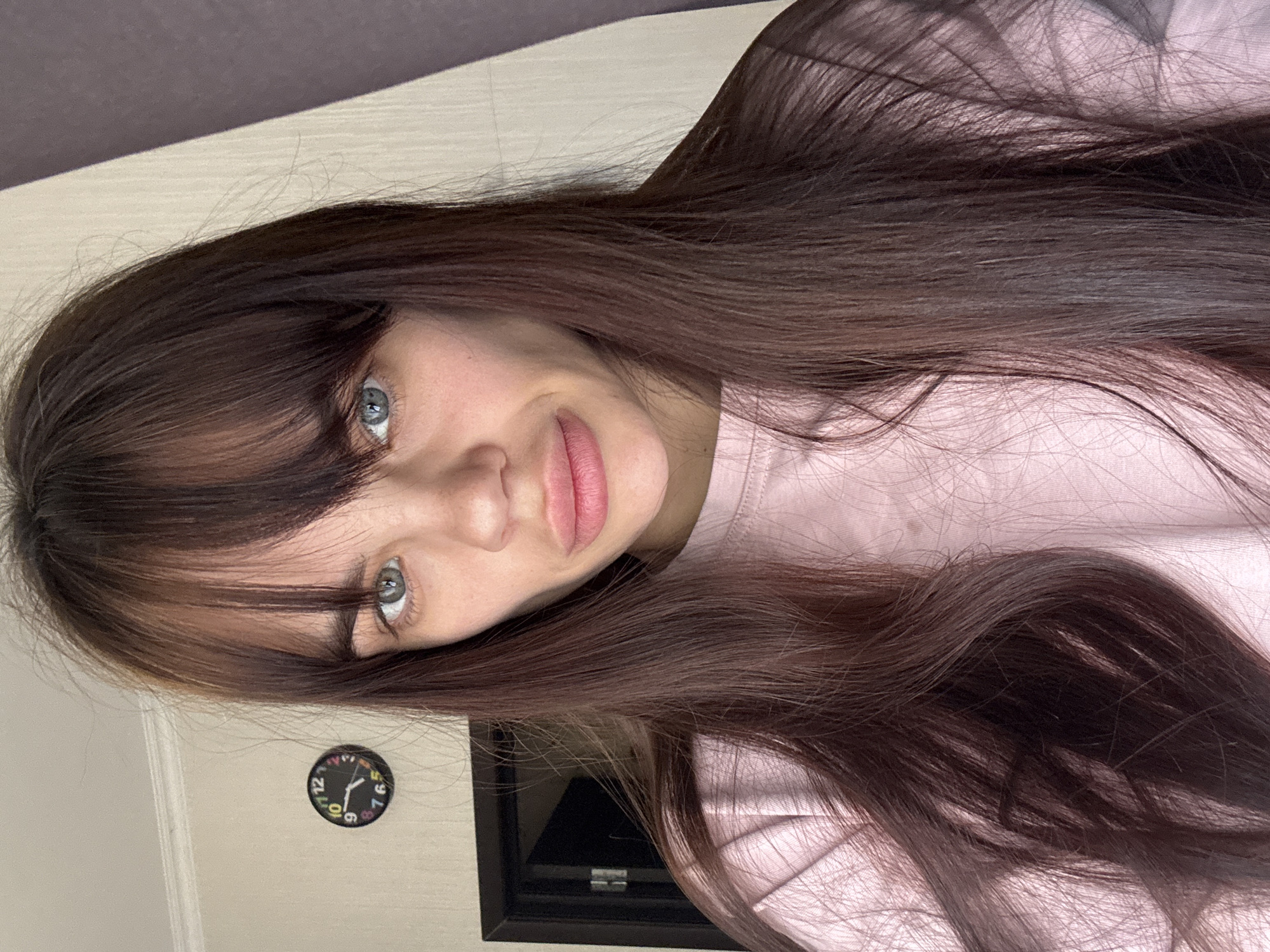 This indoor portrait features a young person, likely female, positioned with her head tilted, suggesting she might be lying down, though the image itself is rotated 90 degrees clockwise. She has long, straight, dark brown hair with full bangs covering her forehead, framing her striking light blue-grey eyes. She looks directly at the camera with a neutral, calm expression. Her lips are full and tinted a natural pink. She is wearing a light pink long-sleeved top or sweatshirt.
In the background, a plain, light-colored wall is visible. To the left side of the frame (when viewed as rotated), a small, round wall clock with a black face and colorful, multi-colored numbers (including 1, 2, 3, 4, 5, 6, 7, 9, 10, 11, 12) is partially seen. The clock hands indicate the time is approximately 9:55. A dark, possibly wooden, frame is also visible in the bottom right corner of the background. The scene is set in an indoor location in Rostov-on-Don, Russia.