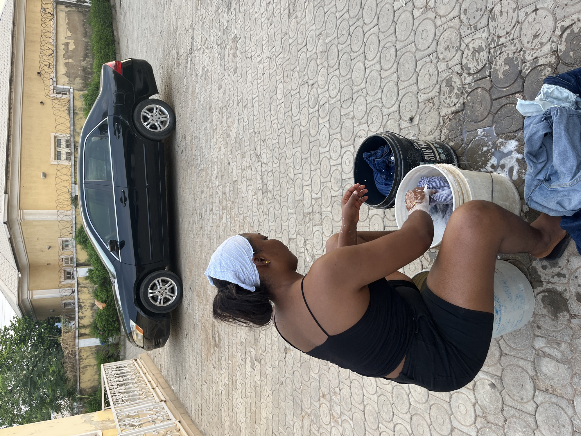 A woman in a black top and shorts is washing clothes in a white bucket, sitting on another bucket in Abuja, Nigeria. There is a black car parked nearby, and a building with barbed wire in the background. The ground is paved with circular patterned tiles. Another bucket with clothes is adjacent to the woman.