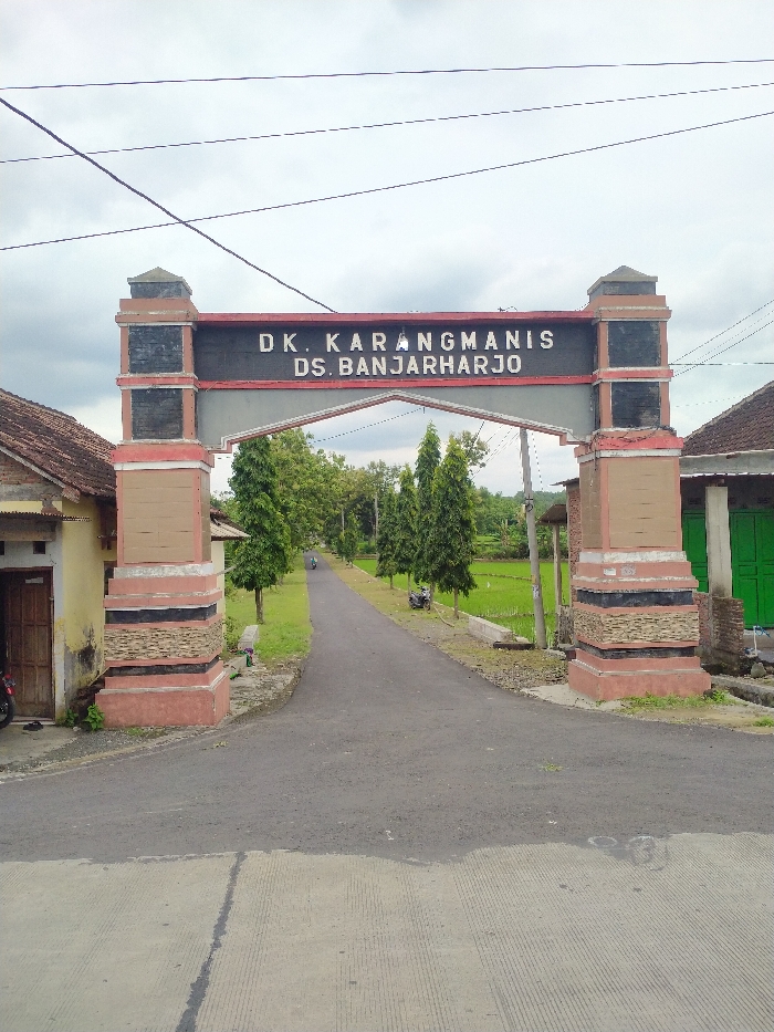 This image depicts the entrance to a rural area in Kecamatan Kebakkramat, Indonesia, characterized by a prominent gate bearing a location sign. The gate features two robust pillars with layered stone-like textures in reddish-brown, black, and beige, supporting a dark horizontal beam. Clearly inscribed on the beam are the words "DK. KARANGMANIS" on the upper line and "DS. BANJARHARJO" below, indicating the Hamlet of Karangmanis within Banjarharjo Village.
A paved asphalt road leads directly through the gate and recedes into the distance, flanked on both sides by tall, slender evergreen trees. On the right side of the road, beyond the trees, lush green rice paddies are visible, signifying an agricultural landscape. On the left, partially obscured by the gate pillar, is a traditional house with a tiled roof. Another house, with a green door, is visible to the right.
In the mid-distance on the road, a person is seen riding a motorcycle, while another motorcycle is parked on the right shoulder. Overhead, several utility wires crisscross the cloudy sky, suggesting an overcast day. The scene conveys a quiet, rural village setting during daytime, with no specific activity beyond the passage of the lone motorcycle.