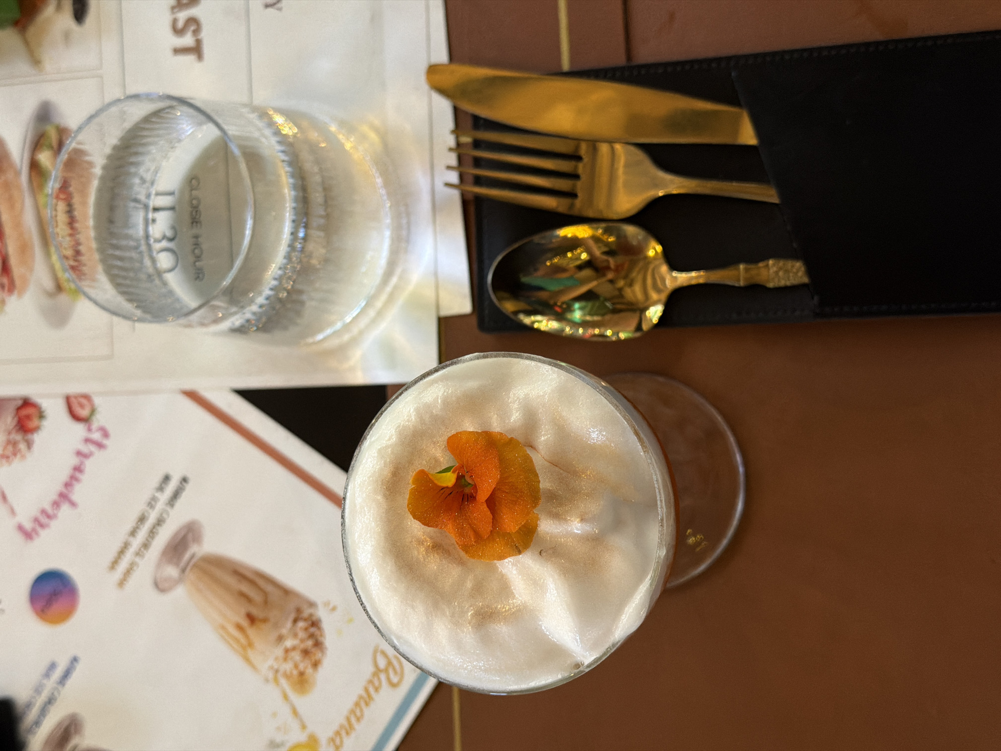 An overhead shot captures a tabletop setting, likely in a cafe or restaurant. The main subject is a sophisticated drink presented in a stemmed glass, topped with a thick layer of white foam or whipped cream. A vibrant orange edible flower serves as a garnish on the foam, which is also dusted with a shimmering golden powder.
To the left of the drink, a clear glass of water stands on the table. The water glass distinctly reflects text that reads "CLOSE HOUR 11.30". Further left, a portion of a menu is visible, featuring illustrations and text. Readable words on the menu include "Strawberry" and "Banana", suggesting options for fruit-based beverages or desserts. A blurry image of a drink on the menu is also discernable, with the text "MILK ICE CREAM SHAKE" partially visible next to it. Other partial text like "AST" (possibly part of "BREAKFAST") can be seen near the top edge of the menu.
To the right of the drink, a set of polished gold-colored cutlery – a knife, fork, and spoon – is neatly arranged in a black, stitched holder. The spoon is highly reflective, showing distorted reflections of colorful objects or lights from the surroundings. The table surface beneath these items is a uniform, warm brown color. The overall scene suggests a pleasant dining experience, with attention to presentation.