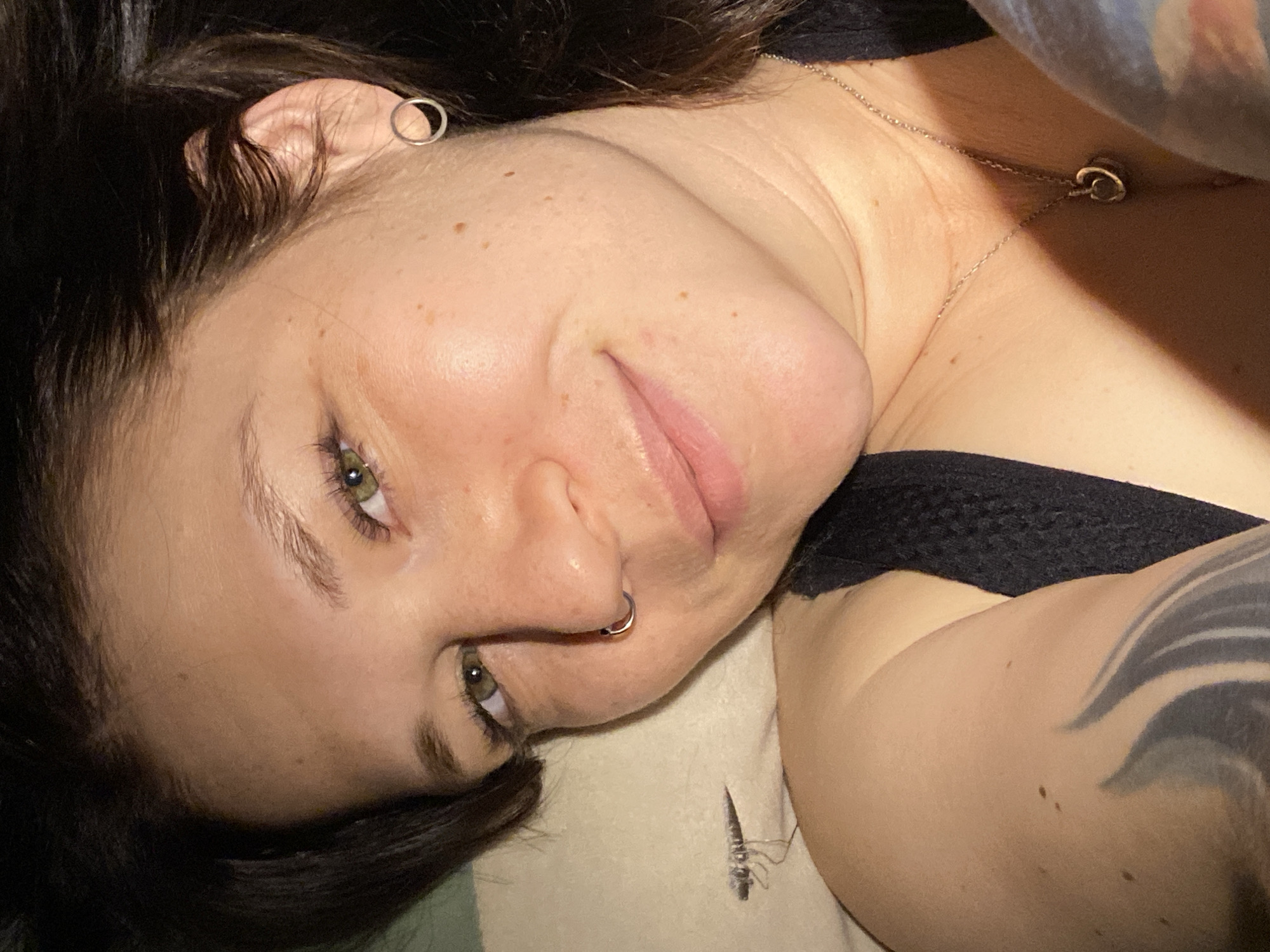 A woman with dark hair and green eyes is lying down, facing the camera. She has a silver hoop earring in her left ear and a silver ring in her left nostril. She is wearing a black tank top and a silver necklace. A tattoo is visible on her upper right arm. The background includes a pillow or soft surface she is resting on. There is nothing in the image to indicate the location of Chaikovsky, Russia.