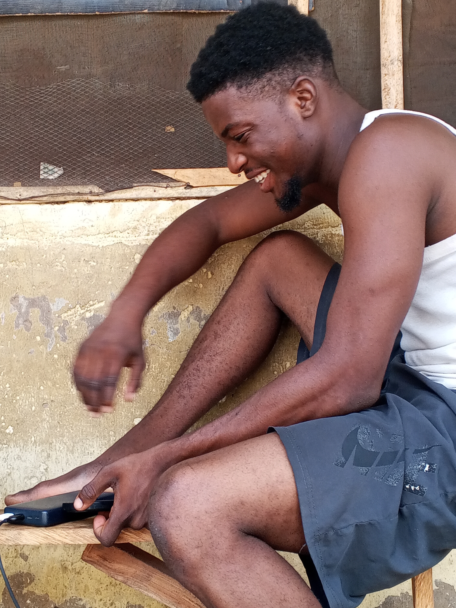 An African man is seated on a wooden surface, likely a stool or bench, located outdoors. He is fair skinned and has an Afro and short beard. He wears a white tank top and black Nike shorts. He is smiling and looking down at a small, dark, rectangular object he is holding in his left hand, which appears to be connected to a charging cable. A wire mesh screen is behind the man on the wall. The environment appears to be a simple outdoor setting with a wall in the background. The location is Lokoja, Nigeria.