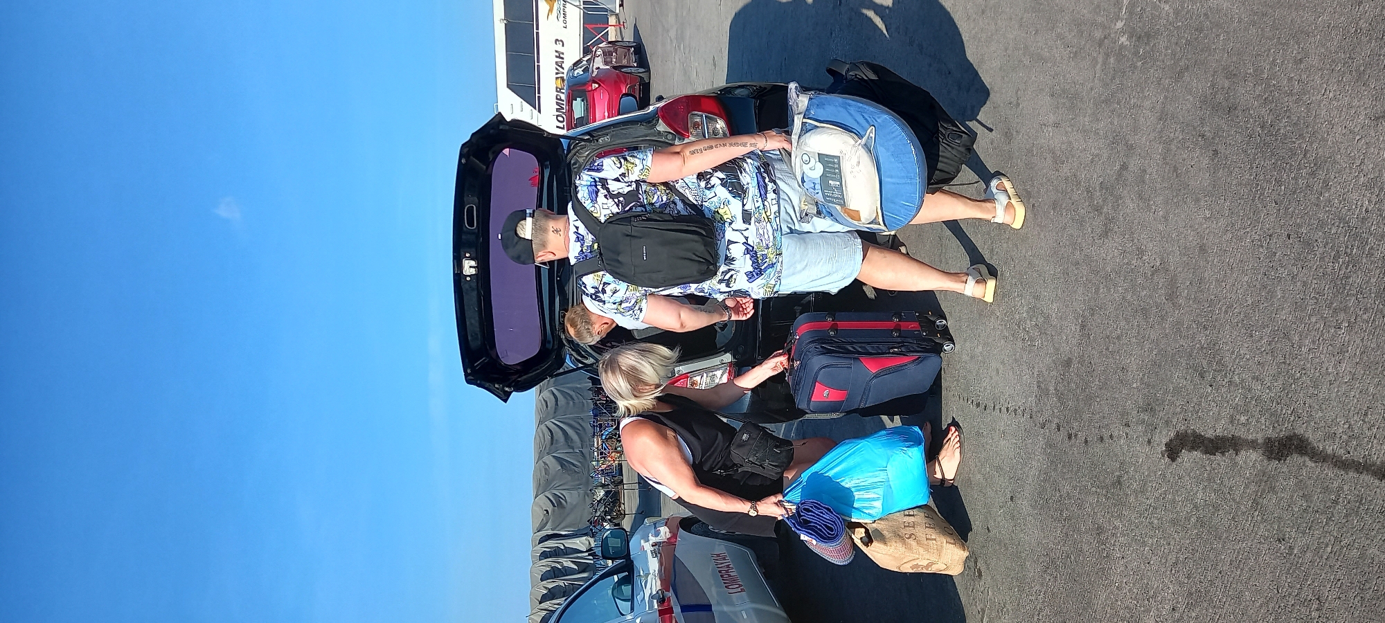 This image depicts an outdoor scene at what appears to be a busy transport hub or ferry terminal in Thong Sala, Thailand, under a clear, bright blue sky. Two individuals are seen actively engaged in loading or unloading luggage from the open trunk of a black car.
On the right side of the car's trunk, a person, likely a man, dressed in a light blue patterned shirt, light-colored shorts, and a black cap, leans into the trunk. He wears a black fanny pack or crossbody bag and white platform sandals. A prominent tattoo on his right forearm reads "ONLY GOD CAN JUDGE ME." Beside him, a large, circular blue package, possibly containing a sleeping mat or bedding with the visible text "NATURA," is placed on the ground next to a black backpack.
On the left side of the trunk, another person, likely a woman with blonde hair, wearing a black top, shorts, and black sandals, is also handling luggage. A blue and red rolling suitcase stands upright nearby. Other items include a bright light blue plastic bag, a blue and white striped woven mat, and a burlap-like bag with partially visible text "SEE FREE."
The ground is asphalt, and the bright sunlight casts distinct shadows, suggesting a clear day. In the background, a large white vehicle, resembling a bus or shuttle, displays "LOMPRAYAH 3" on its side, while another white or silver vehicle in the foreground also has "LOMPRAYAH" visible. These are clear indicators of the Lomprayah ferry company, confirming the setting as a ferry departure or arrival point in Thong Sala. The overall scene portrays travelers managing their belongings, likely transitioning between land transport and a ferry journey.