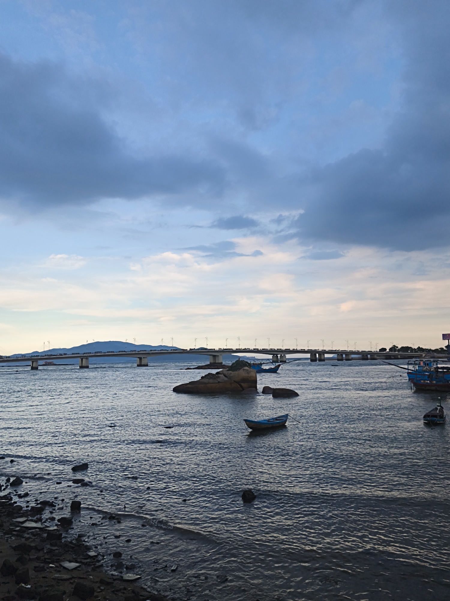 The image depicts a wide, calm body of water in Bac Nha Trang Ward, Vietnam, under a dusky, cloudy sky. A long, multi-span concrete bridge stretches across the water in the mid-ground, with a mountain range visible behind it. The bridge supports streetlights and a traffic lane with visible vehicles. Several small boats are scattered across the water: a blue rowboat is near a cluster of rocks in the center, and two larger blue fishing boats are closer to the right edge of the frame. The foreground shows a rocky shoreline with some dark, wet stones and sand. The overall atmosphere is tranquil, possibly at dusk or dawn.