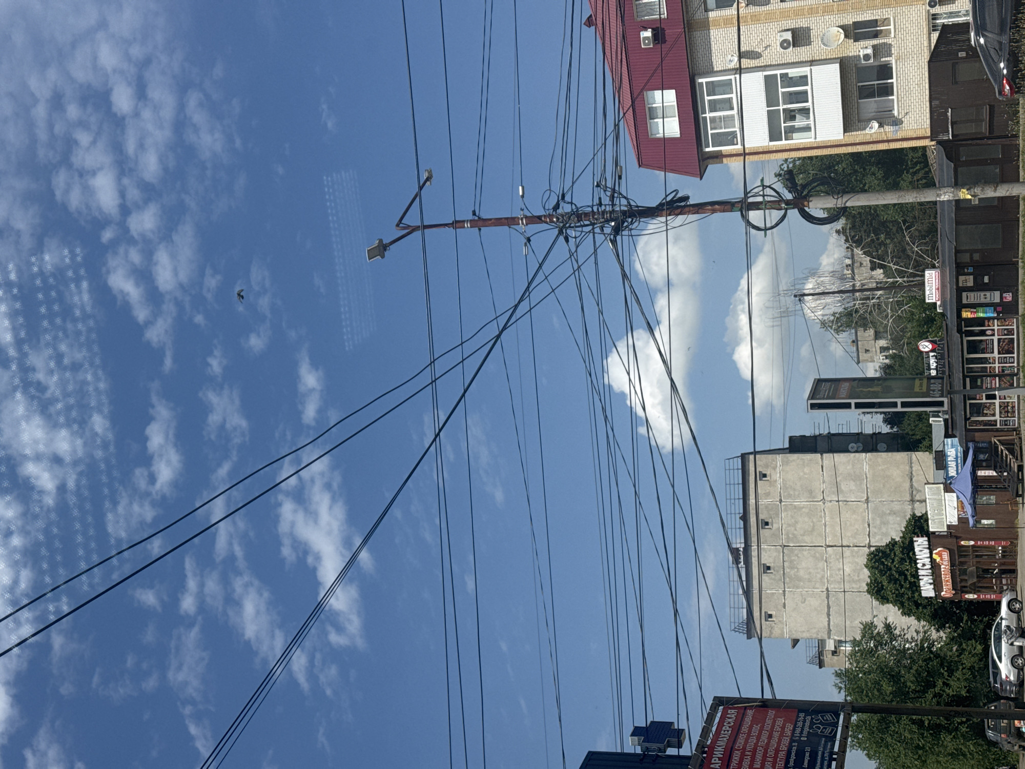 This is a photograph taken on a bright, partly cloudy day in the city of Mikhaylovsk, Russia, likely from a moving vehicle. The composition is dominated by a dense network of overhead electrical wires crisscrossing the sky. In the foreground, a utility pole is heavily laden with these wires, creating a tangled mass of black lines. To the right, a multi-story apartment building with a red tiled roof and light-colored brick facade is visible. It features multiple windows and balconies, with air conditioning units mounted on the exterior. To the left of the utility pole, across a street with parked cars, is a row of low-rise commercial buildings. One building has signage that appears to be in Cyrillic script, and a blue awning is extended over an entrance. Further in the background, a large, grey, Brutalist-style apartment building with a grid-like facade and small square windows is prominent. Trees and other structures are visible behind these main buildings, indicating an urban or suburban setting. A solitary bird is in flight in the upper portion of the sky. The overall impression is of a typical street scene in a Russian city, characterized by functional infrastructure and residential and commercial buildings under a clear blue sky.