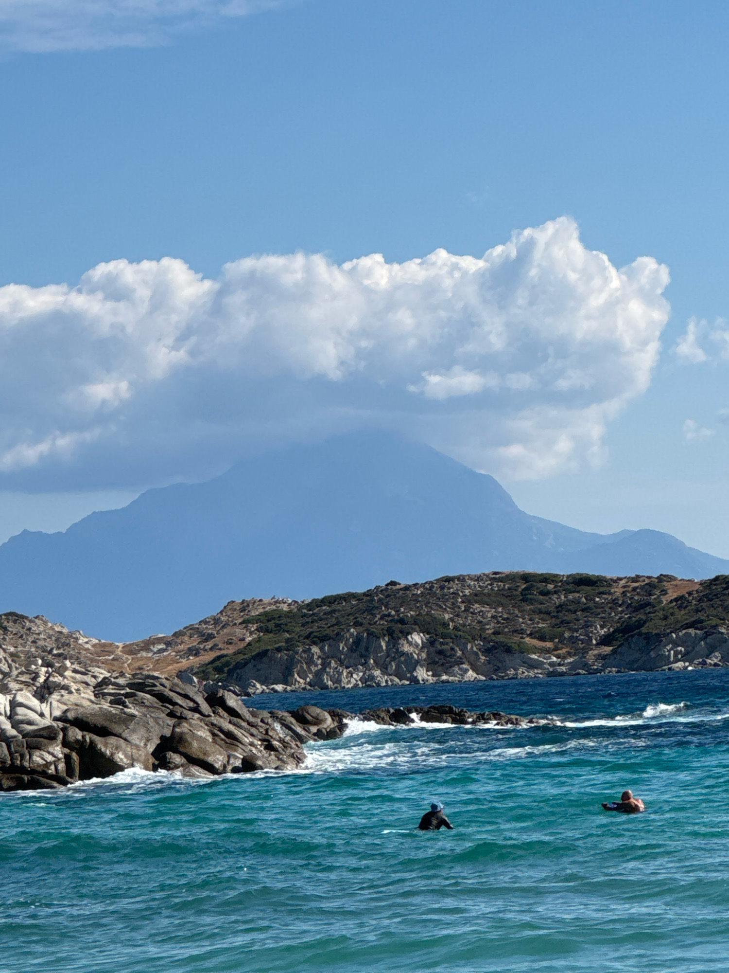 The image captures a coastal scene with a large mountain range in the background, partially obscured by clouds. In the foreground, a rocky shoreline meets the vibrant turquoise water. Two individuals are visible in the water, appearing to be swimming or wading. One person is further out and appears to be floating on their back, while the other is closer to the shore and is facing away from the camera. The sky is a clear blue with scattered white clouds. The scene suggests a day at the beach with recreational water activities.