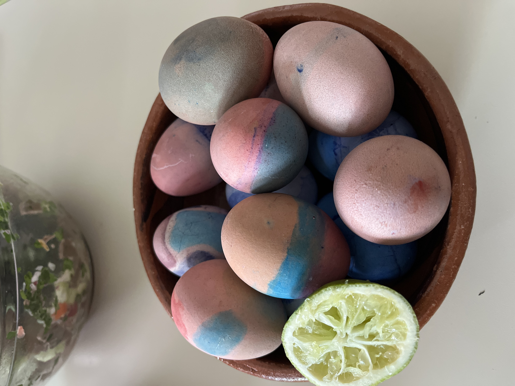 A bowl of dyed eggs and a lime half sit on a white surface. The eggs are dyed in blue and pink hues and fill the brown ceramic bowl. The lime is a light green color. A glass bowl filled with green vegetables can be seen to the left of the bowl of eggs. The location is San Bernardino Tlaxcalancingo, Mexico.