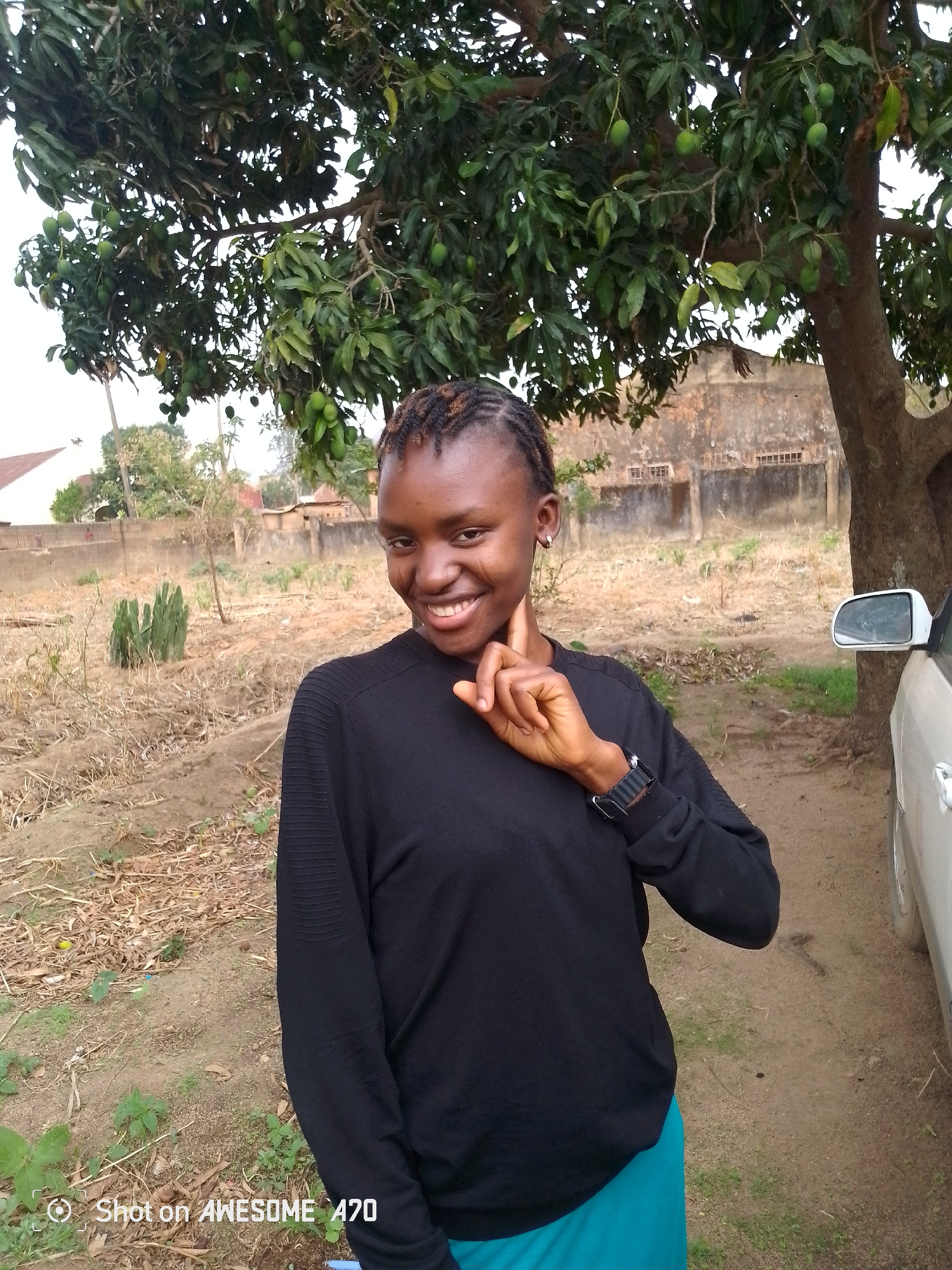 A young person stands outside in the city of Unknown, Unknown. They are smiling and gesturing with one hand near their cheek. They wear a black sweater and teal or turquoise skirt. Behind the person, there is a mango tree, some other vegetation, a dirt ground, and a building. A car is partially visible on the right side of the image. The image resolution is low and a watermark indicating "Shot on AWESOME A70" is at the bottom left.