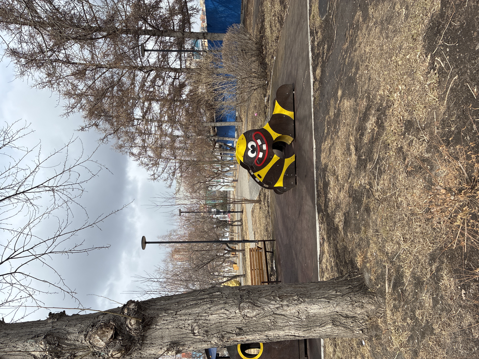 A medium shot captures a children's playground in a park in Unknown, Unknown. Dominating the scene is a black and yellow caterpillar-shaped climbing frame, positioned on a paved path. A wooden park bench sits nearby. The foreground features a tree trunk, extending vertically into the frame. Beyond the climbing frame, evenly spaced lamp posts line the pathway, which is flanked by barren trees and a blue fence in the background. The sky is overcast. Dry grass and withered foliage cover the ground surrounding the path.