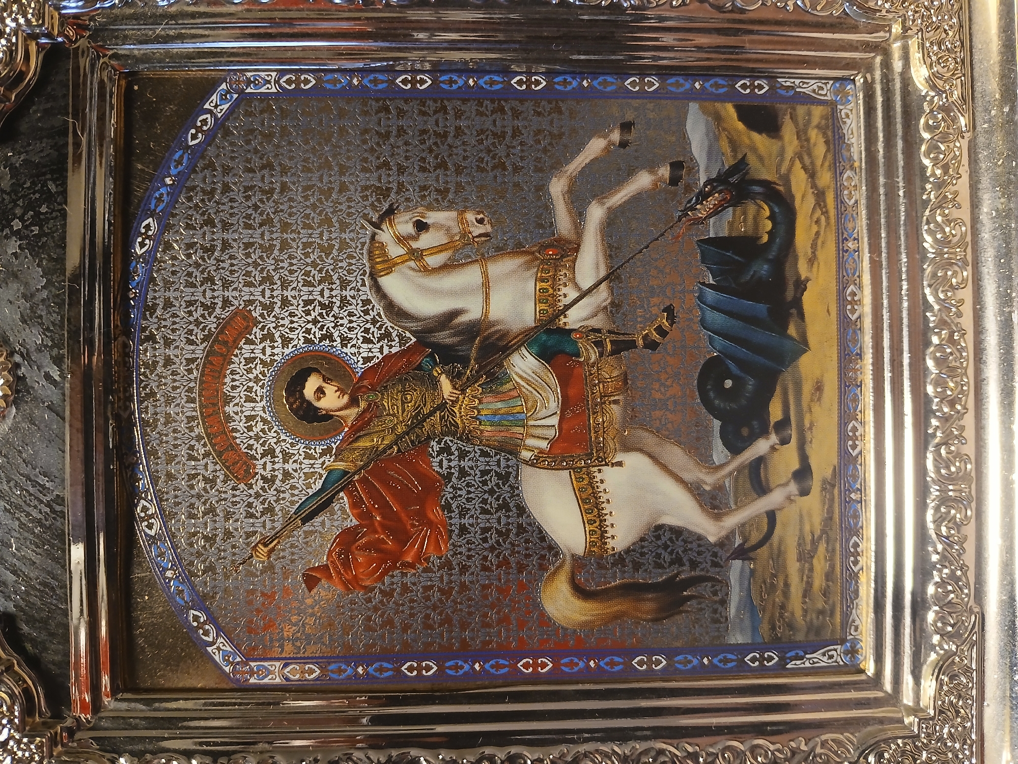 The image shows a framed religious icon depicting Saint George slaying a dragon. The icon is rectangular with a decorated border and a silver frame. Saint George, wearing armor and a red cloak, is mounted on a white horse and is in the act of piercing the dragon with a spear. The dragon is dark blue or black. The background of the icon has a patterned texture. The icon appears to be indoors, based on the lighting and the surrounding environment including a silver decorative border, and is likely located in Kamianske, Ukraine.