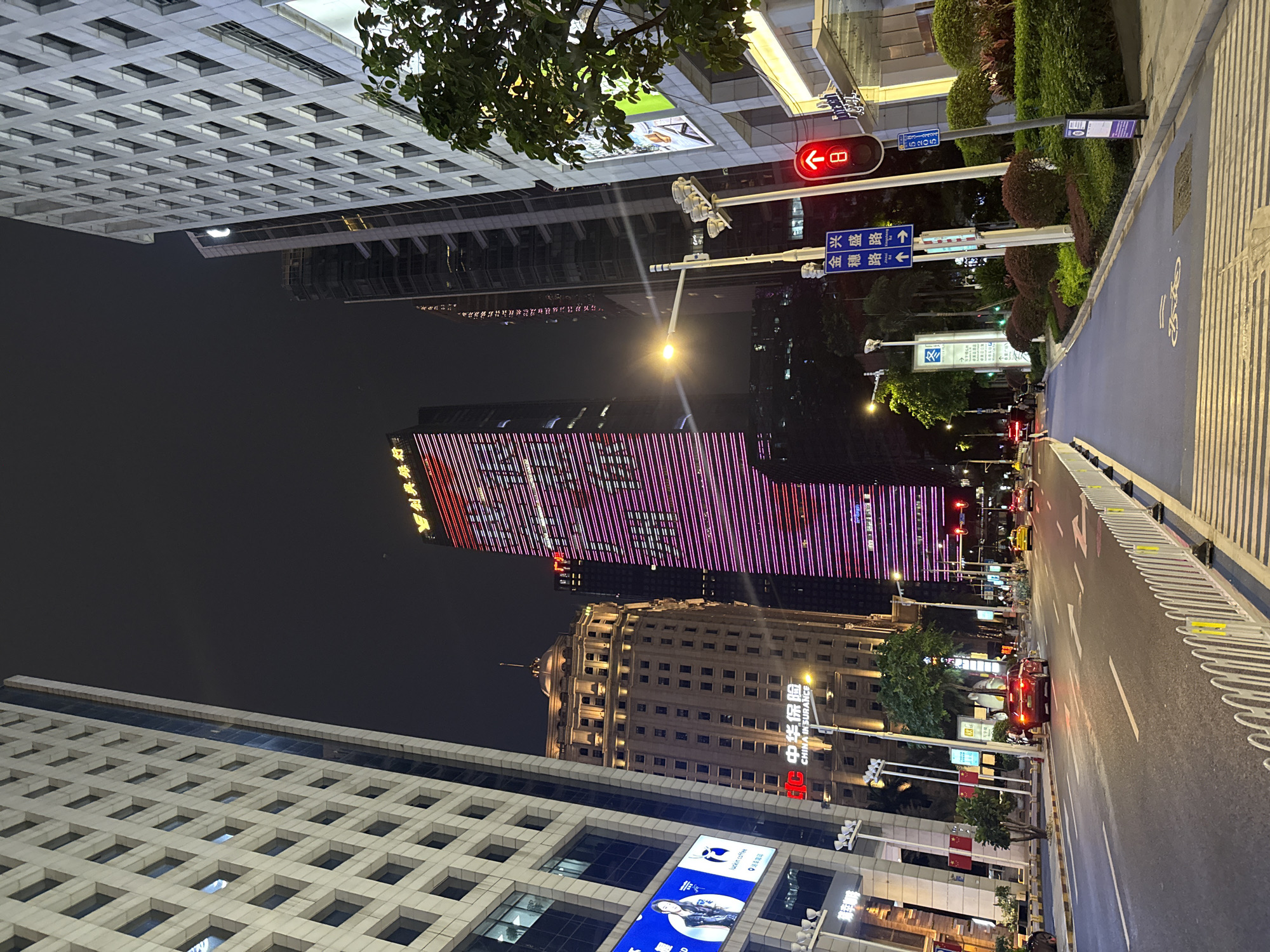 Here is a forensic description of the image:
The image shows a street scene at night in what appears to be a commercial district of Haizhu District, China. Tall buildings line the street on either side. The building on the left has a grid-like facade, with advertisements visible on its lower portion. The central building in the background is lit up with pink and red vertical stripes. Another building next to it displays the characters for "中华保险 CHINA INSURANCE". The street is well-lit, and a blue cycle lane is marked on the right side. Street signs with Chinese characters are visible, including signs for "金穗路 Jinsui Road" and "兴盛路 Xingsheng Road". Traffic lights are also seen with a red light illuminated. A pedestrian walkway runs along the road and vegetation is present. Several cars are visible in the distance, along with other traffic. A streetlight illuminates the buildings.