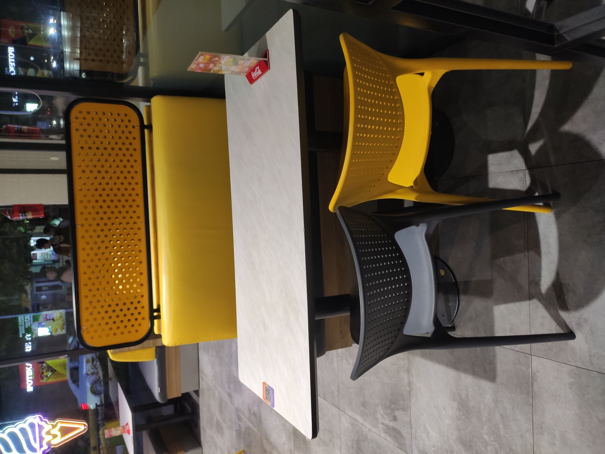 The image captures a seating area within a restaurant, likely a fast-food establishment, in Tashkent, Uzbekistan. The scene is dominated by a bright yellow booth with a perforated backrest and a light-colored, rectangular table in front of it. Two chairs are positioned at the table: one is dark gray and the other is yellow. A small menu or promotional stand, featuring a Coca-Cola logo, sits on the table. In the background, through a window, a street view is visible, with a car, neon signs for "APOTEK" (pharmacy), and indistinct figures of people. The overall atmosphere suggests an indoor dining space at night.