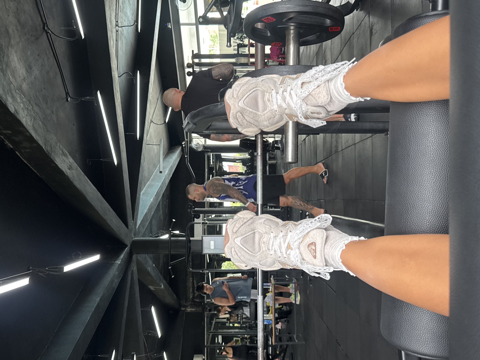 A first-person shot taken in a gym-like setting in Kathu, Thailand. The camera angle is from the point of view of someone sitting on exercise equipment with legs extended and wearing New Balance sneakers. Various people are exercising in the background, including one individual using a weight machine and another walking while wearing flip-flops. The gym has black walls with fluorescent lights running along the ceiling.