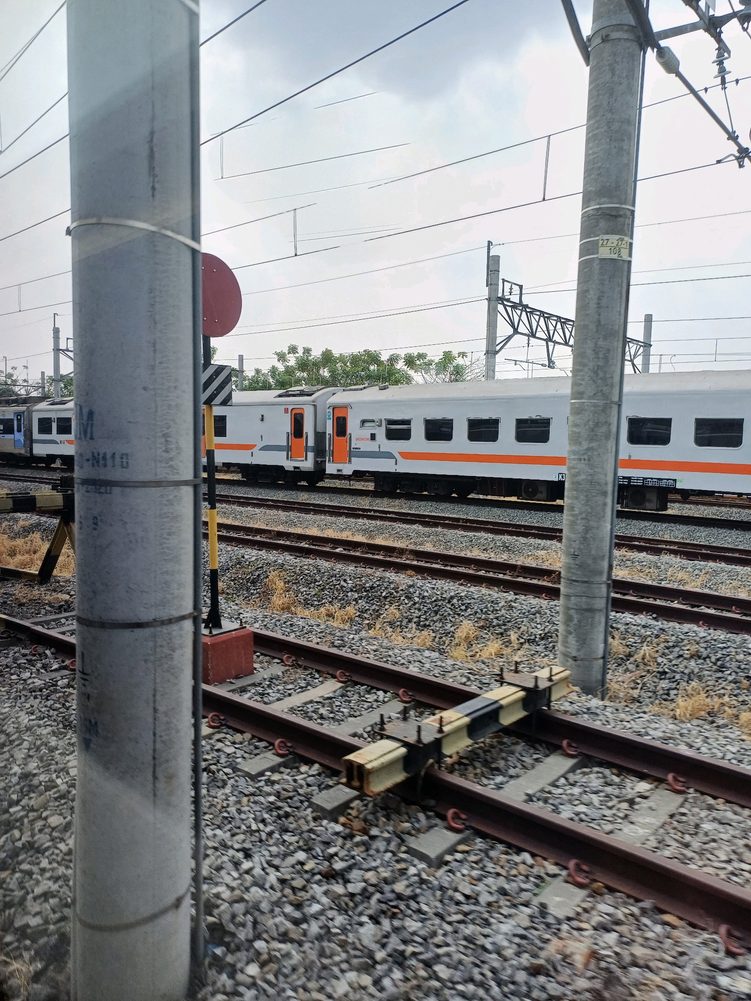 The image depicts a train moving on a railway track in Surakarta, Indonesia. A concrete pole in the foreground obscures part of the view. Behind the pole, a train with white carriages and orange accents is visible on parallel tracks. Overhead power lines crisscross the sky, supported by poles. The ground is covered with gravel and scattered dry vegetation. A railway switch mechanism is prominent in the middle ground. The sky is overcast.