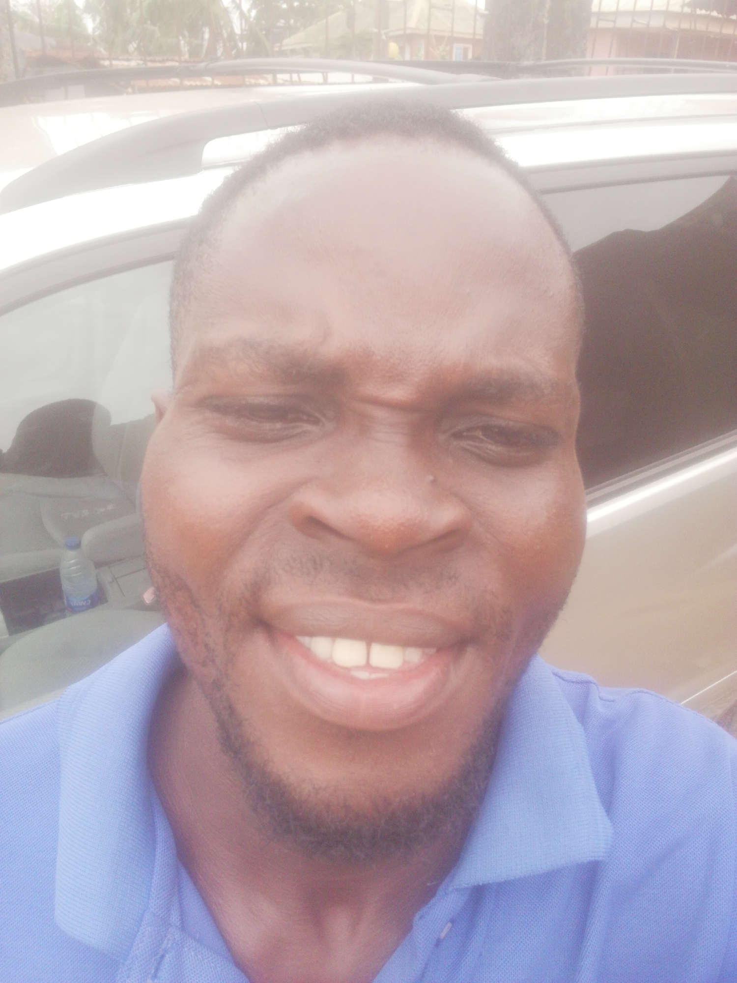A close-up selfie shows a smiling Black man wearing a blue collared shirt. He appears to be inside a vehicle, with a silver roof rack visible above him. A water bottle and interior car details are visible in the background. The location is the city of Unknown, Unknown.