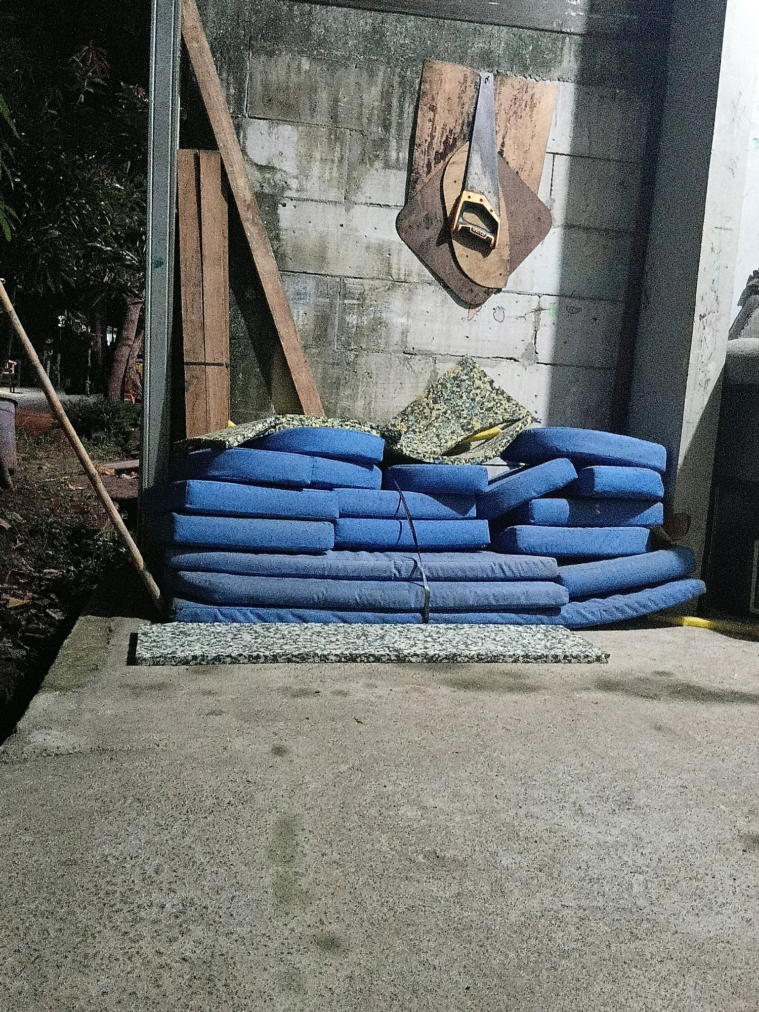 In Cikal, Indonesia, a nighttime scene depicts a stack of blue cushions resting against a concrete block wall. The cushions are of varying sizes and appear to be used for seating. One of the cushions has a black tie wrapped around it. Above the cushions, on the wall, a piece of wood is mounted, with what appears to be a saw blade and handle attached to it, resembling a decorative or functional item. To the left of the cushions, there are two wooden planks leaning against a metal beam. The ground in front of the cushions is made of concrete. A piece of patterned foam matting is placed on the concrete in front of the stacked cushions. The environment suggests an outdoor area, possibly a backyard or a storage space. The lighting indicates it is dark, with artificial light sources illuminating the scene.