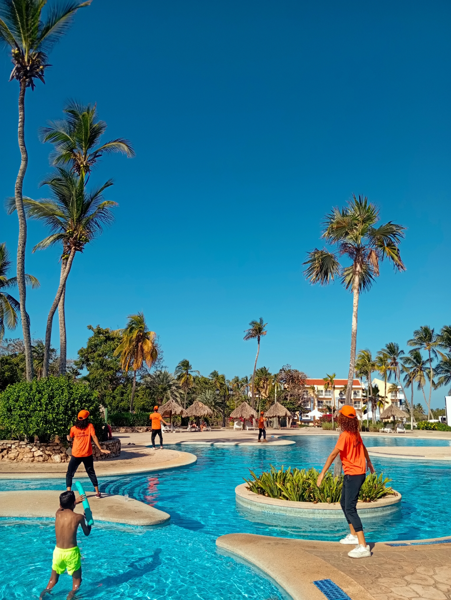 The image shows an outdoor swimming pool area under a bright blue sky. Tall palm trees dot the landscape. Four adults wearing orange T-shirts and hats are positioned around the pool. One adult is standing on a walkway beside the pool, while another is on a concrete island. A child stands in the water holding a blue object. The pool has multiple levels and sections. In the background are thatched-roof structures and a multi-story building. The location is consistent with a resort setting in Playa El Agua, Venezuela.