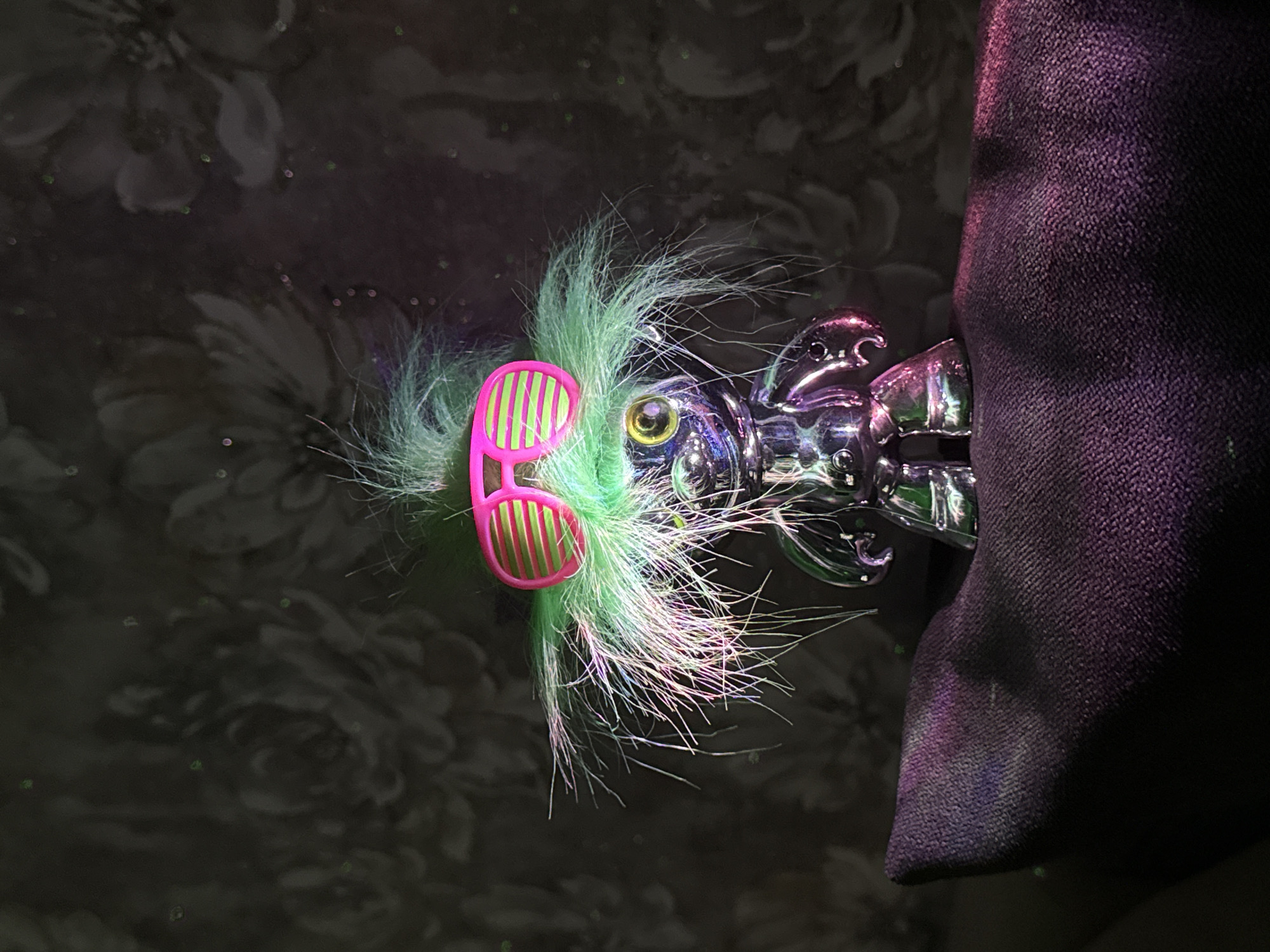 The image shows a novelty item, appearing to be a toy or decoration, positioned against a dark floral background. The item has a metallic body, possibly chromed plastic or metal, and a tuft of green and white faux fur or hair affixed to the head area. The faux fur is adorned with a pair of small, pink sunglasses with green stripes on the lenses. To the right, a section of dark purple fabric is visible, suggesting a larger object or background element. The scene's environment is dark, and the location is not identifiable.