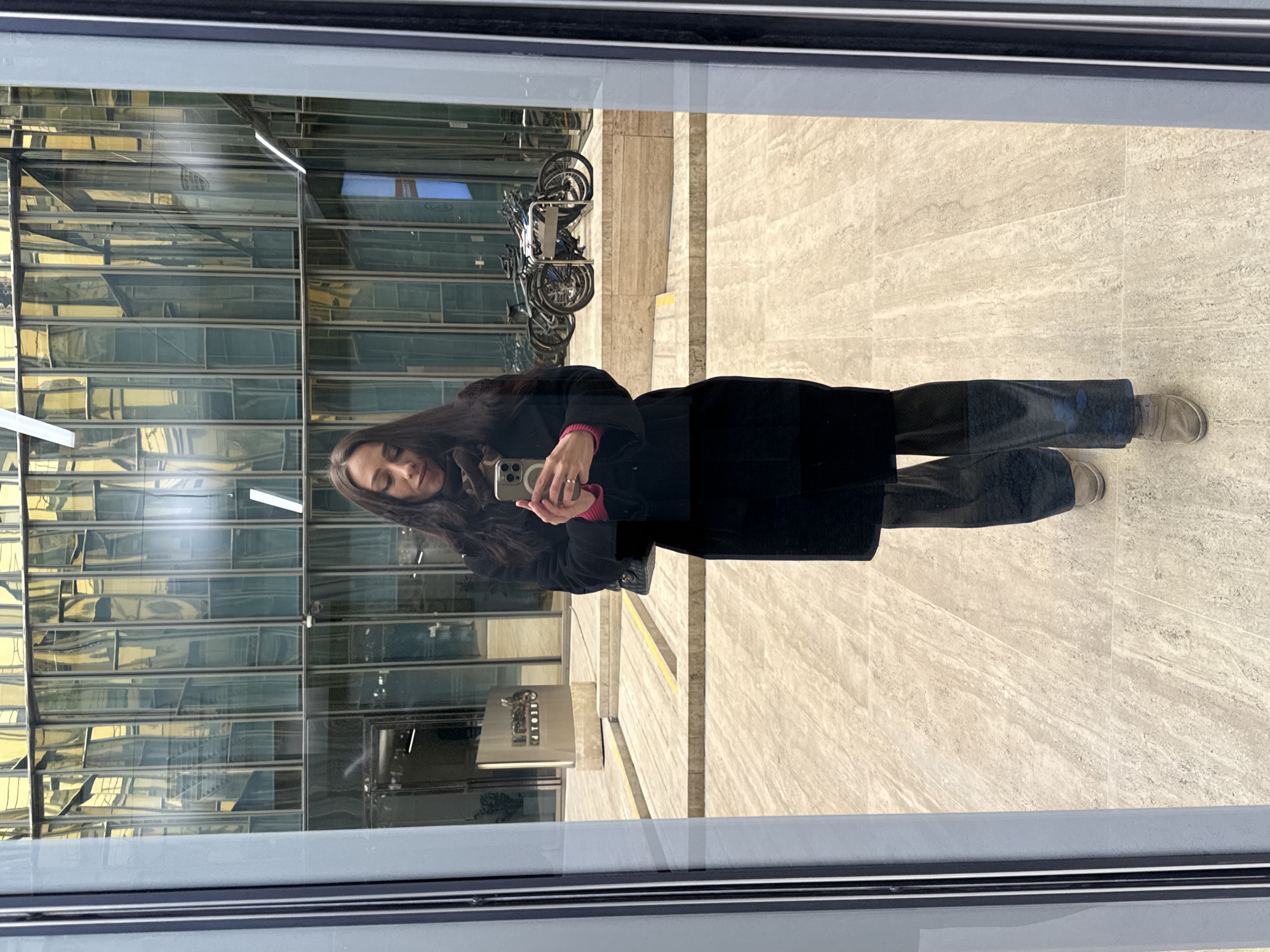 A woman stands in front of a glass window, taking a selfie with a smartphone. Her dark hair contrasts with her light skin. She wears a long black coat over a pink top and dark pants. The reflection in the window shows a modern building with glass windows, a row of bicycles parked outside, and a sign displaying "RESTOR". The floor is tiled with a light-colored stone pattern. Based on the building architecture and the "RESTOR" signage, the location is likely in Saint Petersburg, Russia.