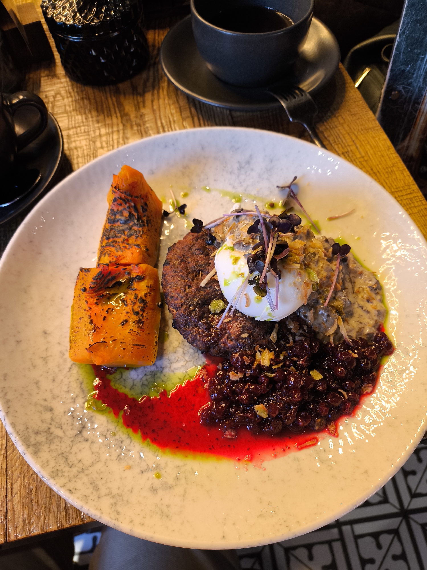 The image presents a high-angle, close-up view of a gourmet meal plated on a round, light-grey speckled ceramic plate, situated in Izhevsk, Russia. The dish comprises a dark, seared patty, possibly a meat cutlet or vegetable burger, topped with a soft-boiled or poached egg. The egg is garnished with purple microgreens and light-colored crispy flakes. Adjacent to the patty is a dollop of a creamy, greyish-brown purée, potentially mushroom-based. To the left of the main components are two elongated, roasted orange vegetable pieces, exhibiting visible charring on their skins, consistent with pumpkin or sweet potato. A glossy, deep crimson berry compote or sauce, likely lingonberry or cranberry, forms a pool at the bottom-right of the plate, bleeding slightly into green oil drizzles that pattern the plate. The plate rests on a textured, light-brown wooden tabletop. In the background, to the upper left, a black ceramic mug with a saucer is partially visible, and another similar black mug and saucer appear to the upper right. A dark, ornate glass container is also partially visible on the table. A black two-tined fork is positioned to the right of the plate. The bottom-right corner reveals a partial view of black and white patterned floor tiles. No individuals are present in the scene, which depicts a static food presentation.