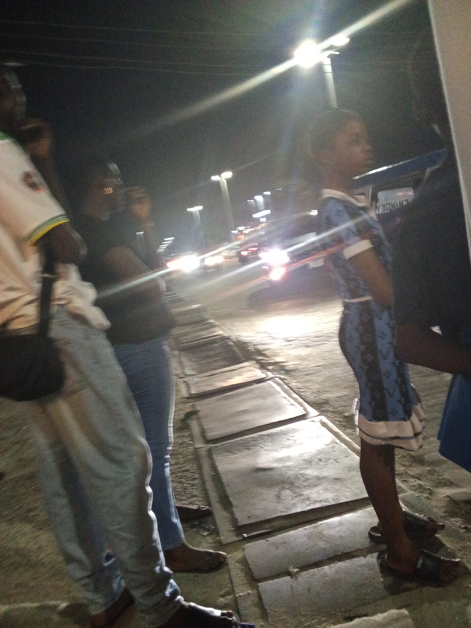 The image depicts a nighttime street scene in Ado, Nigeria, featuring several people standing on a sidewalk alongside a road. The scene is heavily illuminated by bright streetlights and vehicle headlights, creating significant lens flares and strong contrasts that cast some figures into silhouette while others are partially visible.
In the left foreground, a person is seen wearing a light-colored long-sleeved shirt, possibly white with green and yellow stripes on the sleeve, and light blue jeans. They have a black shoulder bag. Adjacent to them, another individual stands in a dark top and light blue jeans, with arms crossed. Further right, a young person, possibly a child or young adult, is visible in a blue dress featuring a white and dark patterned design, looking towards the left. A portion of another person in dark attire is seen on the far right. The individuals appear to be congregating or waiting by the roadside.
In the background, multiple bright vehicle headlights indicate ongoing traffic on the road. The ground consists of large, flat paving slabs, which reflect the light, possibly suggesting a damp surface. Dark lines resembling overhead power lines are visible against the night sky. The setting is characteristic of an active urban or semi-urban environment after dark.
Faint, reversed, and partially obscured text is visible on the back of the blue dress, but it is too blurry and distorted to be definitively deciphered.