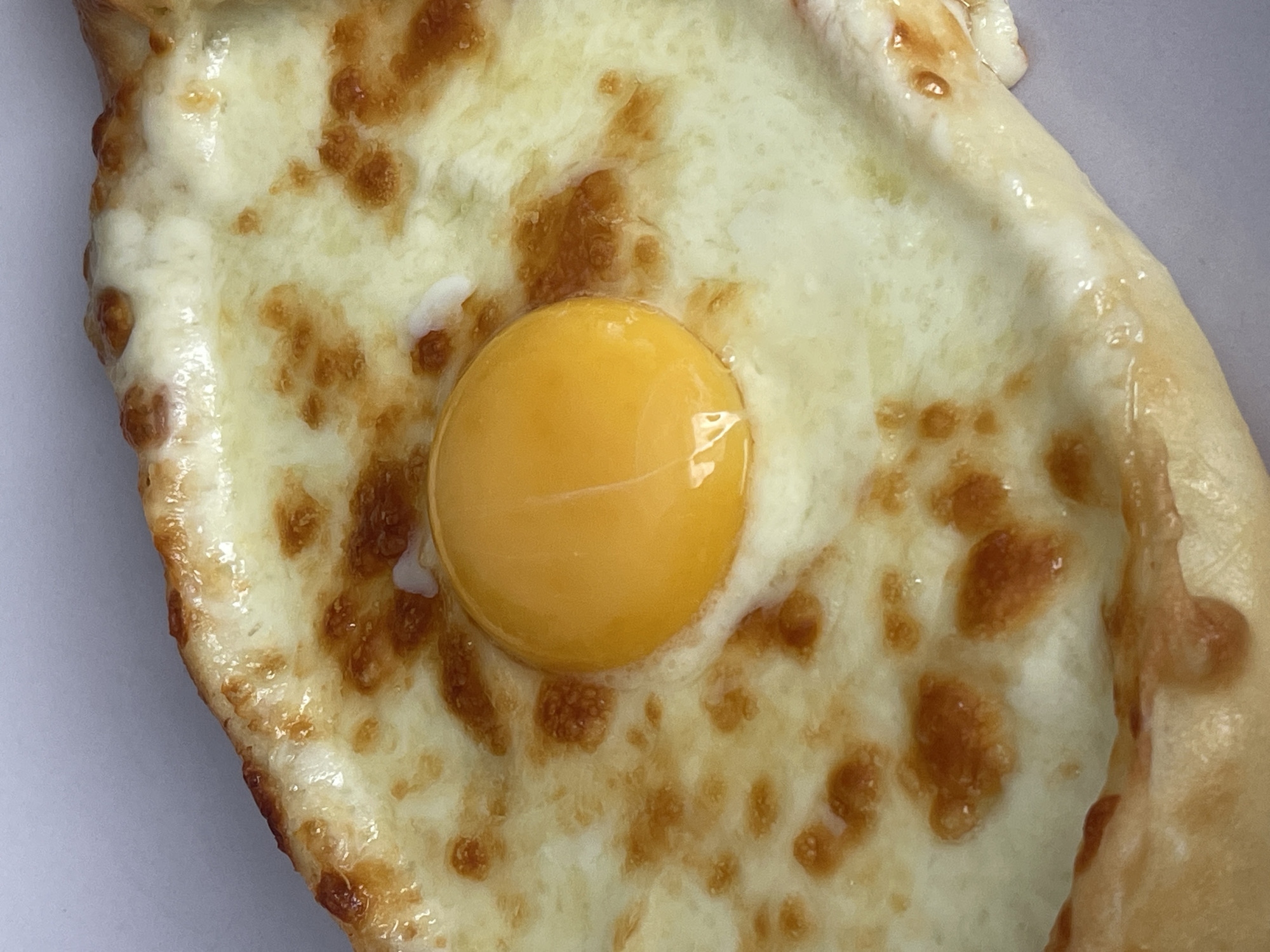 The image is a close-up, overhead shot of a khachapuri, a Georgian cheese-filled bread, served on a light-colored surface, possibly a plate or table, against a neutral background. The khachapuri is boat-shaped, with a thick, golden-brown crust that is slightly charred in places, indicating it was baked. The interior is generously filled with melted, stringy white cheese that has browned in spots, and in the center, a whole, uncooked egg yolk sits prominently, surrounded by a halo of melted egg white. The lighting highlights the glossy texture of the cheese and the vibrant yellow of the yolk. This dish is not specifically located in Orenburg, Russia.