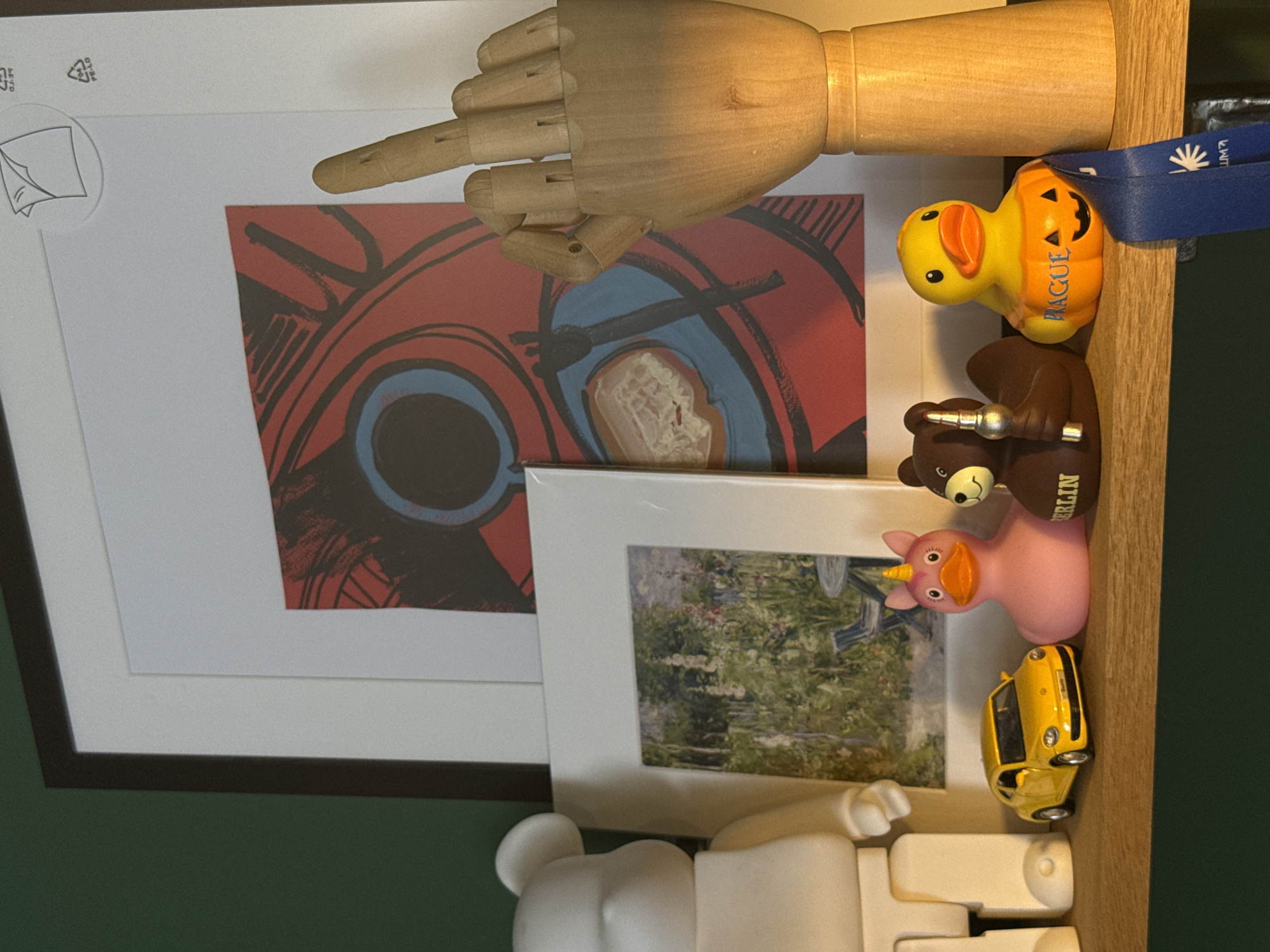 The image shows a collection of objects on a shelf. A wooden artist's mannequin hand is positioned in the upper part of the frame, with its index finger pointing left. Below the hand, there are several decorative items. A framed print with a red and black abstract design is visible, partially obscured by a white mat. Next to it is another framed print featuring a garden scene with flowers.
On the shelf, a yellow rubber duck with a pumpkin design is placed behind a brown bear figurine. The duck has the word "PRAGUE" printed on its side. The bear figurine has a silver object in its paw and the word "BERLIN" printed on its side. A small yellow toy car is parked in front of the garden print. At the bottom of the frame, a white abstract sculpture, possibly a toy, is visible. A blue ribbon with white text is draped over the edge of the shelf near the rubber duck. The background consists of a dark green surface. The lighting suggests it's indoors, likely daytime. There are no people or animals visible.