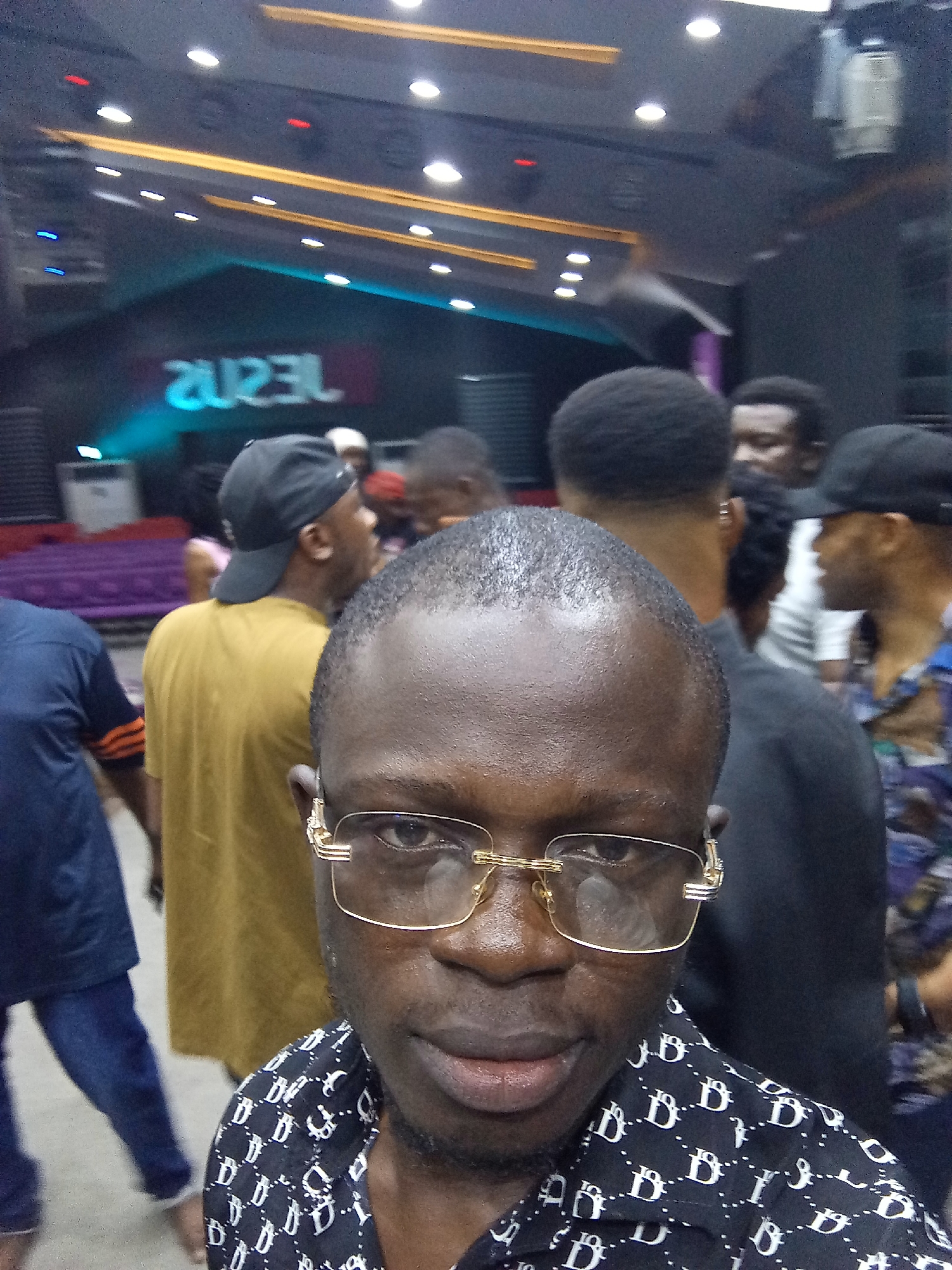 The photo depicts a medium shot of a person with glasses in a black and white patterned shirt, possibly a selfie, taken indoors in Abuja, Nigeria. Several other people are visible in the background. A stage with neon lighting can be seen in the background, displaying a reversed text that reads "JESUS." The location appears to be a performance venue or church.