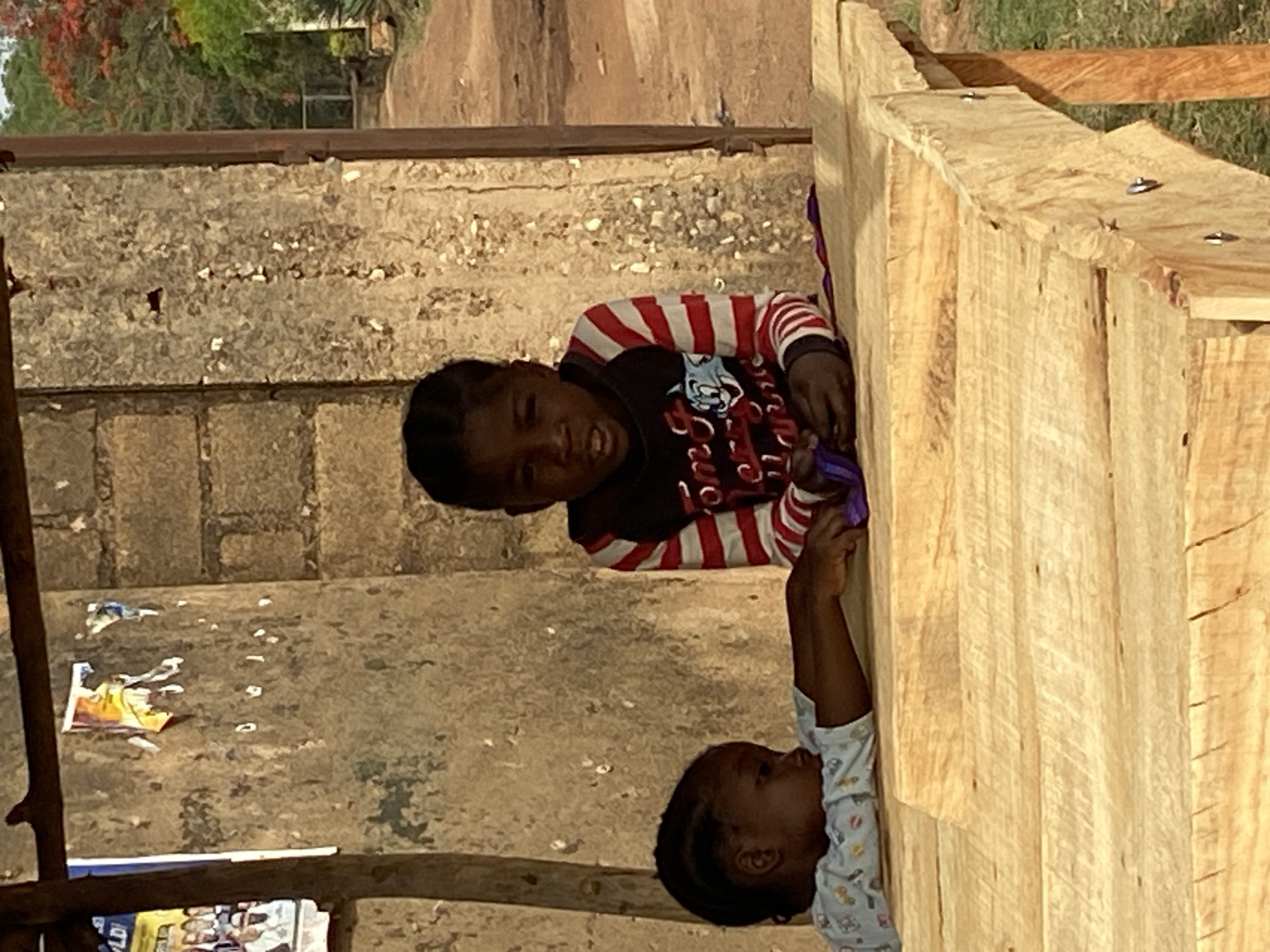 Two children are visible in an outdoor setting. The child on the left wears a red and white striped shirt, while the child on the bottom wears a light-colored patterned shirt. Both children appear to be interacting with a light-colored wooden structure, possibly a building under construction or a play structure. The background shows a weathered concrete structure and a blurred landscape with trees. Based on the prompt, the location is Bukuru, Nigeria.