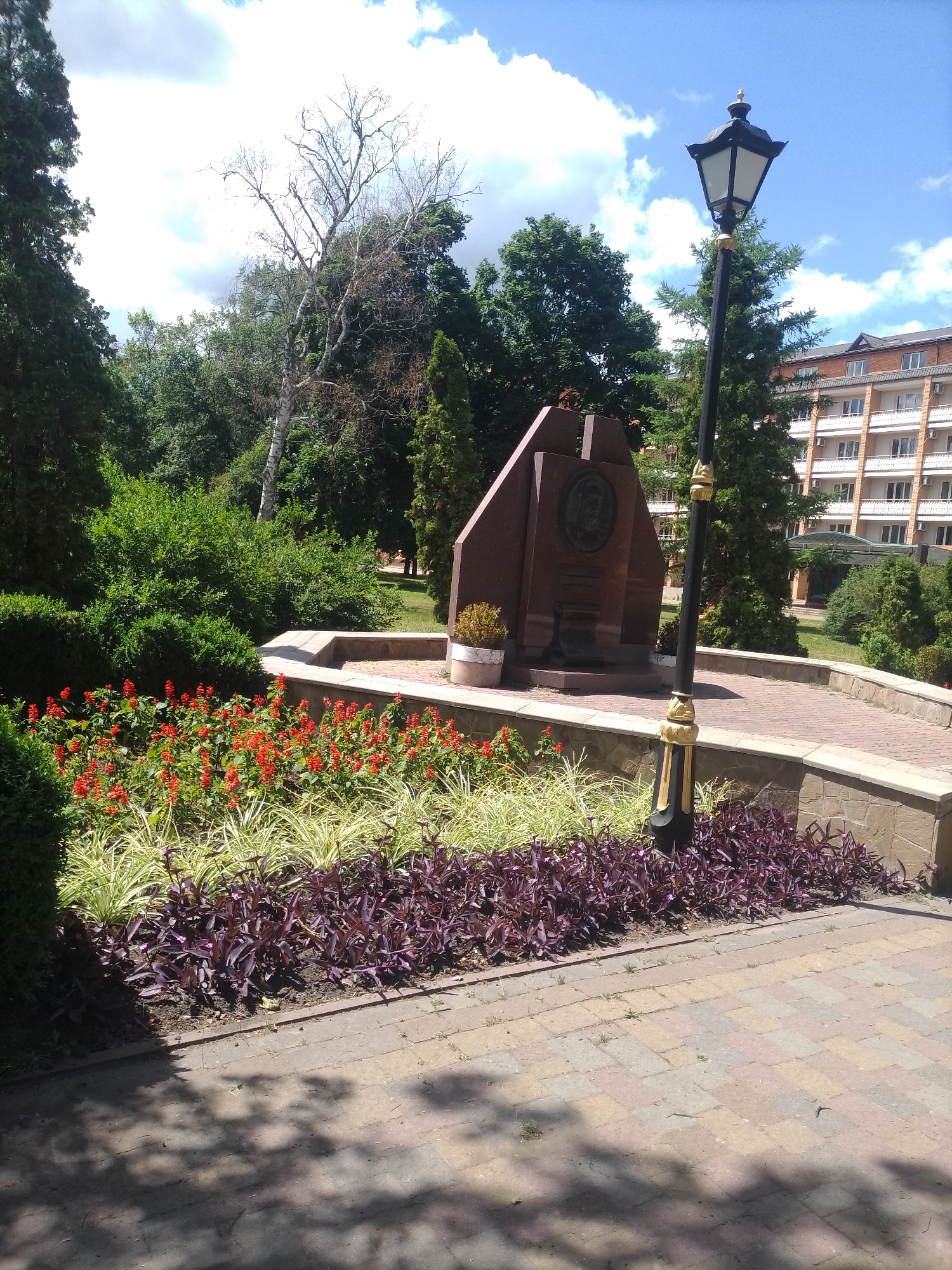 In Myrhorod, Ukraine, a public park area features a stone monument with a circular medallion, possibly commemorating a historical figure. Adjacent to the monument is a vintage-style black lamppost with gold accents. The scene is landscaped with a variety of plants, including a bed of red flowers, a row of light-green foliage, and a border of dark purple leaves. The walkway is paved with brick-like stones, and shadows from surrounding trees are cast upon it. In the background, a multi-story building with a light-colored facade and balconies is visible, as are lush green trees under a blue sky with scattered clouds.