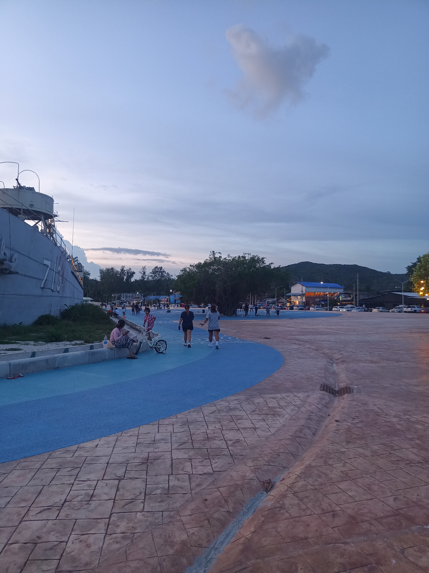 The image depicts an outdoor scene at dusk, likely in a coastal area, given the presence of a large ship and a wide open promenade. The sky is a pale blue with some scattered clouds, suggesting the transition from day to night.
In the foreground, a paved area with a textured, cobblestone-like pattern curves into a bright blue surface. This blue area appears to be a pathway or recreational space, possibly a running track or a dedicated area for leisure activities. The transition between the two surfaces is sharp and deliberate.
To the left of the frame, a substantial grey ship with the number "715" visible on its hull is docked. The ship is partially overgrown with grass and vegetation along its base. A low concrete barrier separates the ship from the blue promenade.
Several individuals are present, engaged in various activities. A woman is seated on the concrete barrier near the ship, seemingly resting or using her phone. Next to her is a small white bicycle. Further along the blue pathway, two people are walking, one wearing dark shorts and a dark top, the other in a grey t-shirt and shorts. Several other figures are scattered in the mid-ground, walking and standing, suggesting a public gathering space.
In the background, lush green trees line the edge of the scene. Beyond the trees, there are some buildings, including one with a blue roof illuminated by lights, indicating commercial activity. A mountain range forms the backdrop, its slopes dark against the twilight sky. Cars are parked in an area between the promenade and the buildings. The overall impression is of a public park or waterfront area in a tropical or subtropical location, possibly a tourist destination, given the amenities and the diverse crowd. The location is identified as Koh Phangan, Thailand, which is known for its beaches and natural beauty, making this scene plausible for such a place.