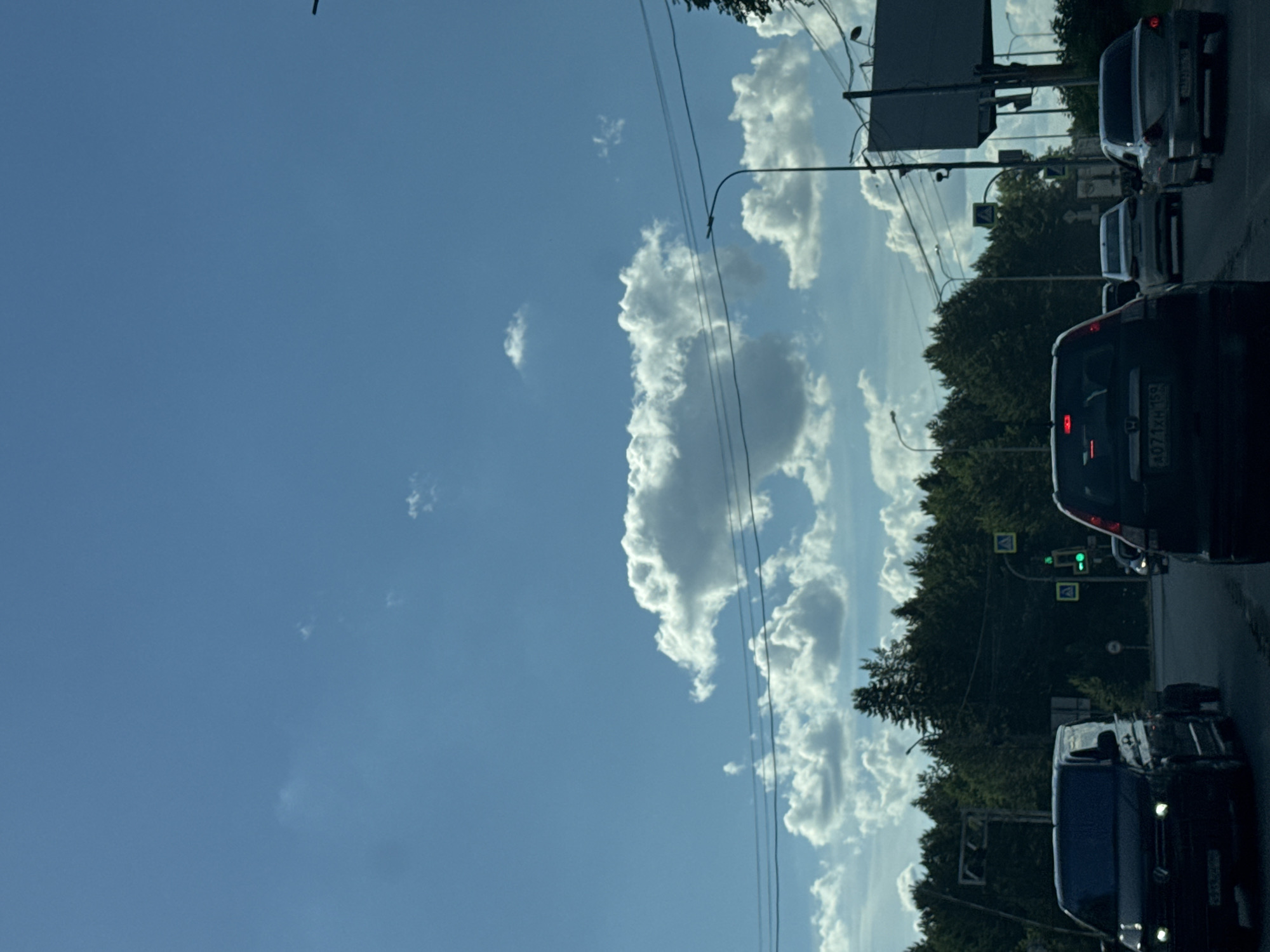 The image, taken in Chaikovsky, Russia, depicts a traffic scene on a sunny day with scattered clouds. The perspective is from inside a vehicle, looking forward. Several cars are visible in traffic, queuing on a paved road. One car's license plate reads "A071XH 159". In the background, lush green trees line the road, and a traffic light displaying a green signal is visible. Overhead, power lines stretch across the frame, and a tall street lamp stands on the right side of the road. The sky is a clear blue with fluffy white clouds, suggesting pleasant weather conditions. The overall impression is of a typical daytime commute or journey.