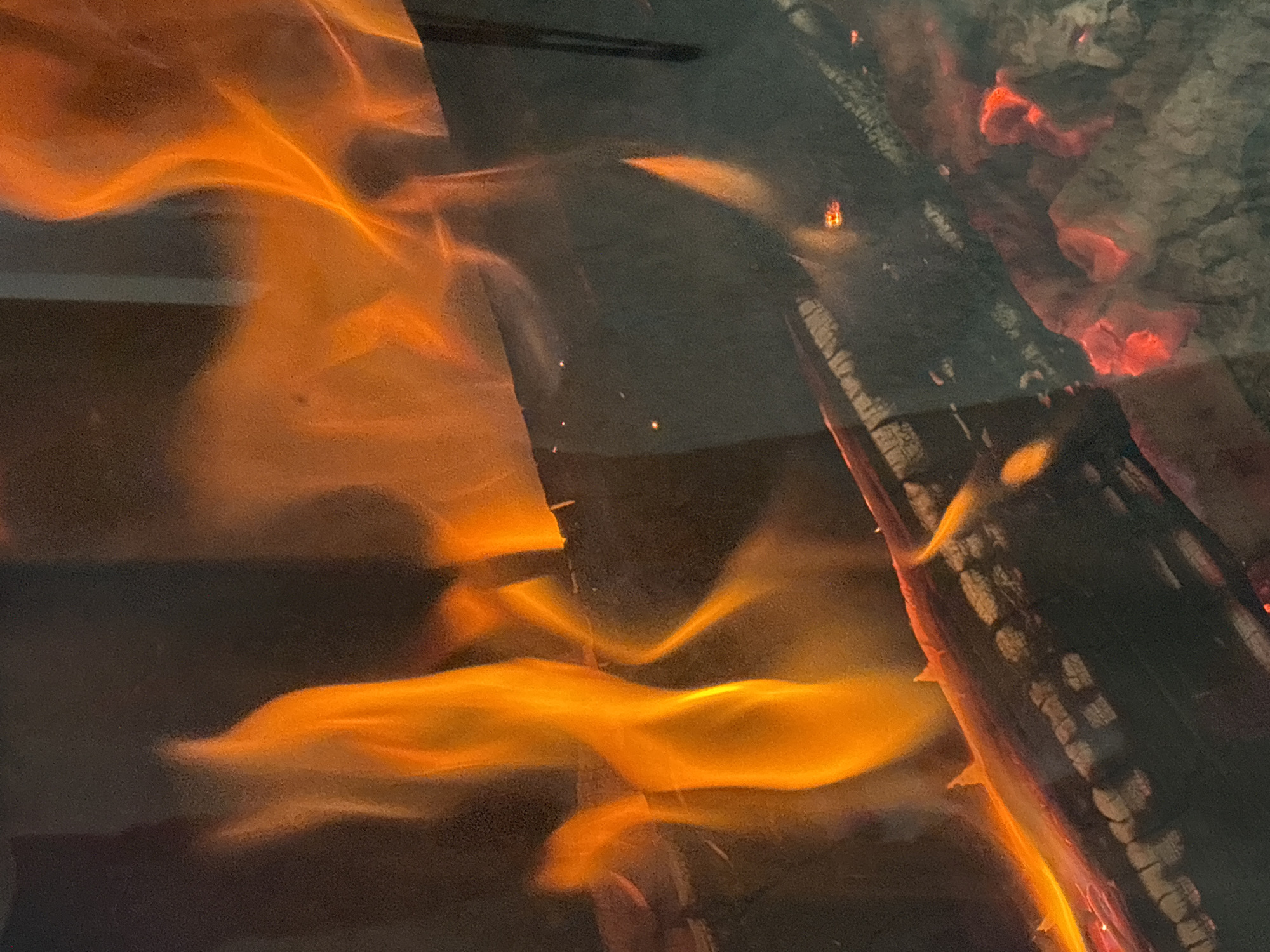 The image shows a close-up view of a fire burning in a fireplace. Several logs are visible, some partially consumed by flames, with glowing embers at the bottom. Bright orange flames lick upwards and around the logs. The background is dark and appears to be the interior of the fireplace or a darkened room. There are no people or animals in the image. The scene is consistent with a domestic fireplace, potentially in a residential setting in Zvenigorod, Russia, given the context provided.