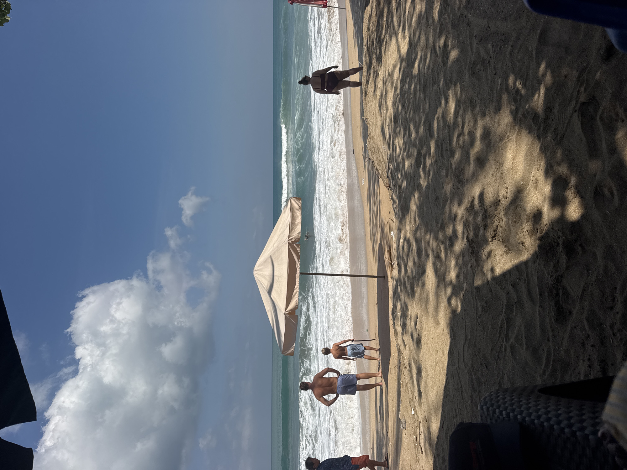 The image shows a beach scene in Kuta, Indonesia. The sun is shining brightly, indicating it is daytime. The sky is clear blue with a few fluffy white clouds. The sand on the beach is light brown and textured with footprints and shadows from palm trees. The ocean is a vibrant turquoise, with moderate waves breaking on the shore.
Several people are visible on the beach. In the foreground, three men are standing on the sand, facing the ocean. One man in the center is wearing blue swim trunks and has his hands on his hips. To his left, another man in blue swim trunks is turned away from the camera, looking towards the water. Further to the right, a third man is partially visible, also in swim trunks.
Further down the beach, closer to the water, a person is standing. In the distance, a large beach umbrella provides shade, and a few other figures can be seen near the water's edge. The overall mood appears relaxed and summery, typical of a beach day. There is no visible text in the image.