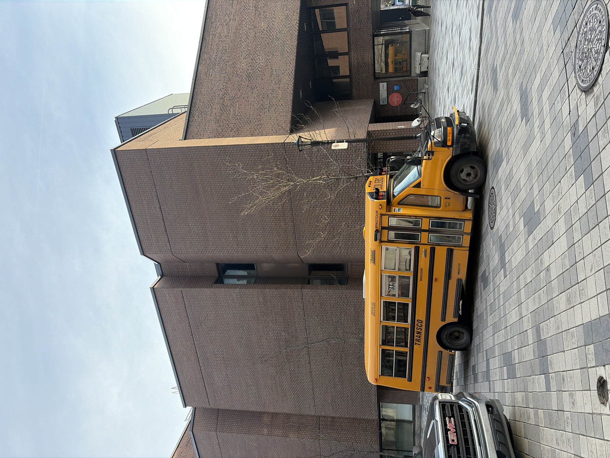 A yellow school bus is parked on a gray and white brick street next to a large, brown brick building. A silver GMC vehicle is partially visible behind the bus. The building appears to be a commercial or institutional building, possibly in downtown Montreal, Canada. A bare tree stands near the bus and the building. The sky is partly cloudy. A manhole cover is visible in the top right corner of the image and another is at the bottom right, which includes lettering "VILLE DE MONTREAL".
