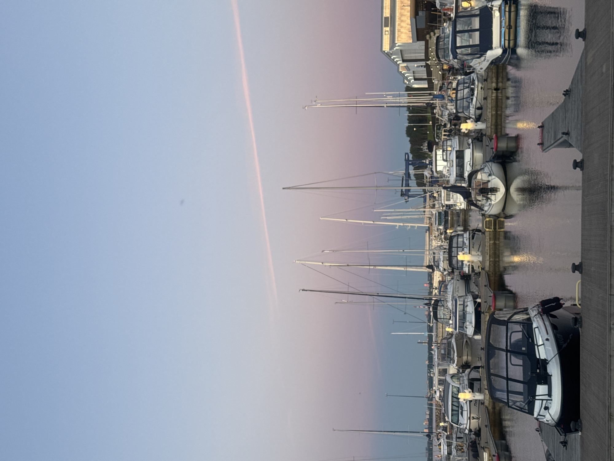 The image depicts a marina in Tallinn, Estonia, during twilight. The sky transitions from a pale blue at the top to soft pink and purple hues near the horizon, with a faint pink contrail stretching across the upper portion. The water of the marina is calm, reflecting the sky and the numerous boats docked along wooden piers.
The foreground features a wooden pier with bollards. A black and white motorboat, partially covered, is prominently docked in the center foreground. Further into the marina, a collection of various sailboats and motorboats are moored. Some sailboats have tall masts with rigging visible, while motorboats vary in size and design, many with covered cabins.
In the background, a modern building with large windows and a light-colored facade is visible along the shoreline, suggesting it might be a boat club or service facility. Trees line the edge of the marina behind the buildings. The overall atmosphere is peaceful and serene, characteristic of a marina at the end of the day. No people are visible in the image.