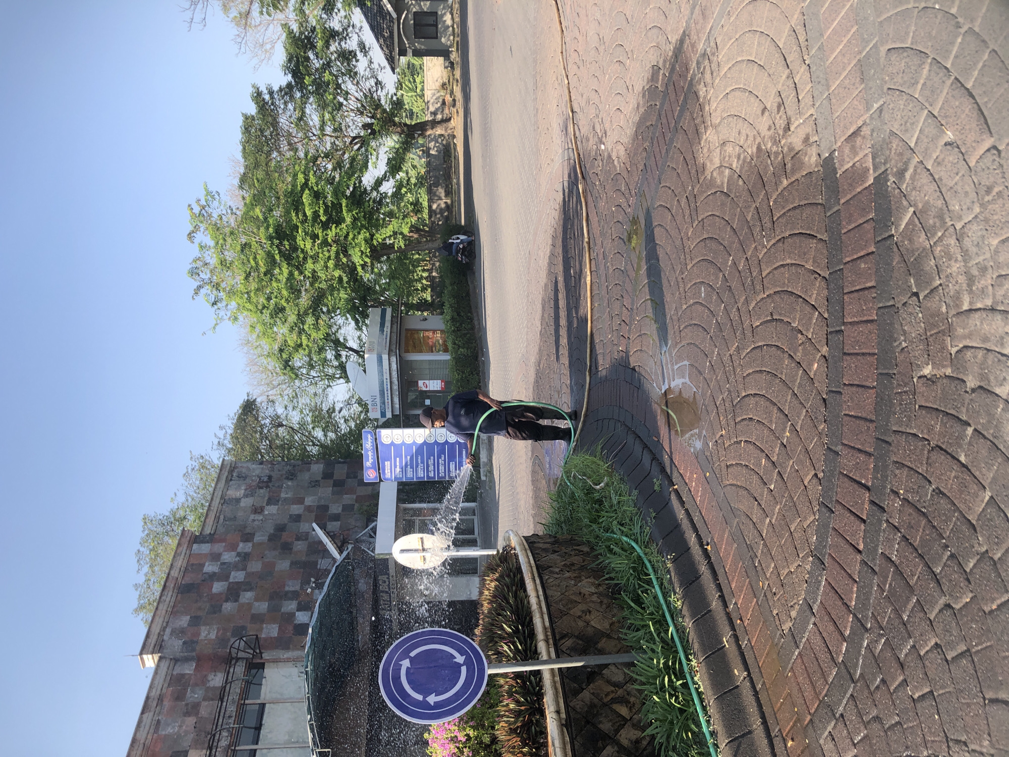 A man is watering plants with a green hose. He is wearing a dark blue t-shirt and dark pants. The man is standing on a brick-paved area. Behind him, there is a circular sign with three arrows indicating a roundabout. There are also some plants and a stone wall. In the background, there is a building with a patterned facade, trees, and a clear blue sky. The scene is taking place outdoors in what appears to be a public space, possibly in Boro, Indonesia given the context. The lighting suggests it is daytime, with the sun casting shadows on the ground.