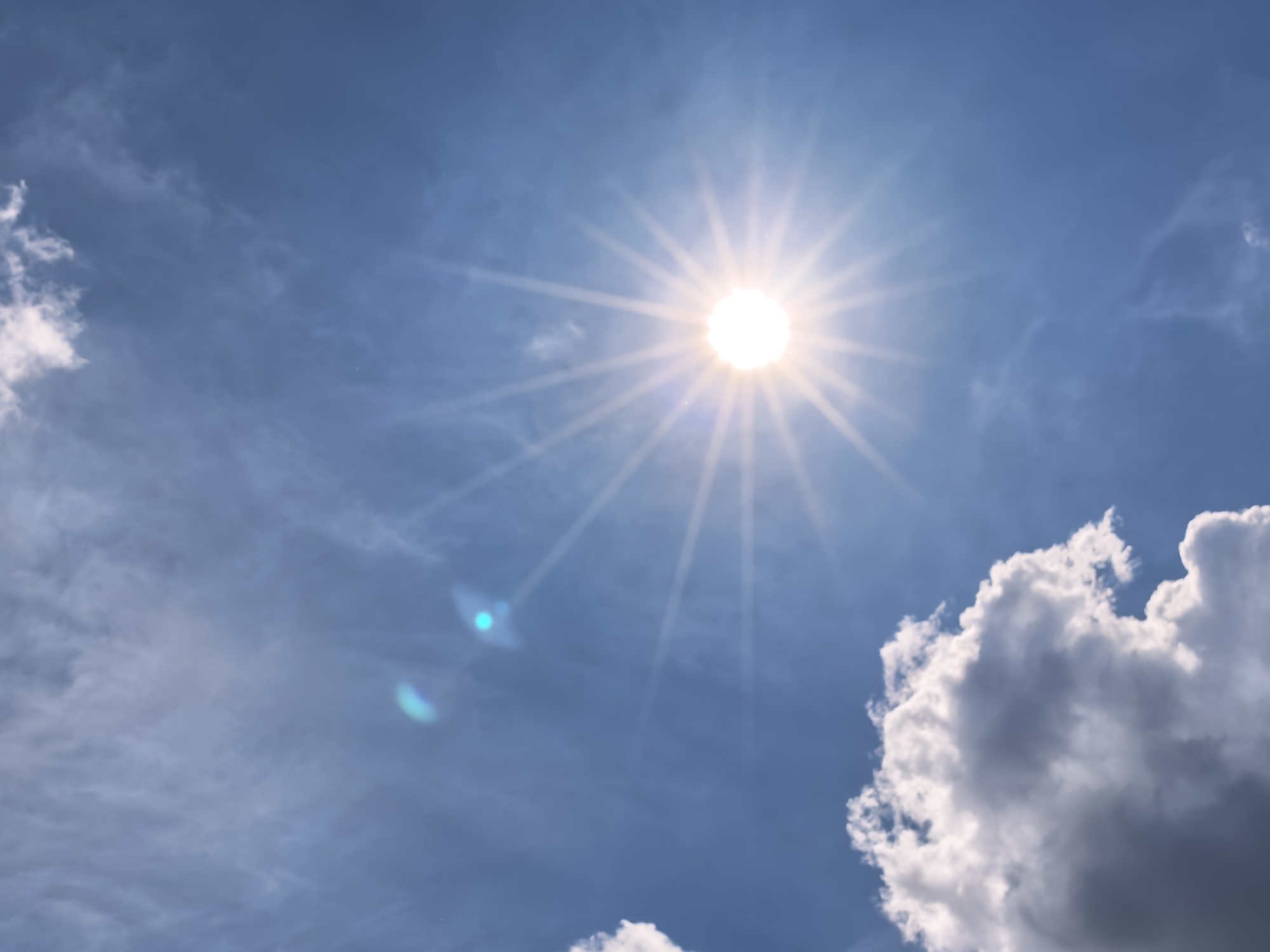 The image depicts a bright, sunny sky with the sun positioned at the center-top. Light clouds are scattered throughout the blue sky, with a larger, fluffy white cloud dominating the lower-right portion of the image. A lens flare is visible in the lower-left. The location is Unknown, Unknown.