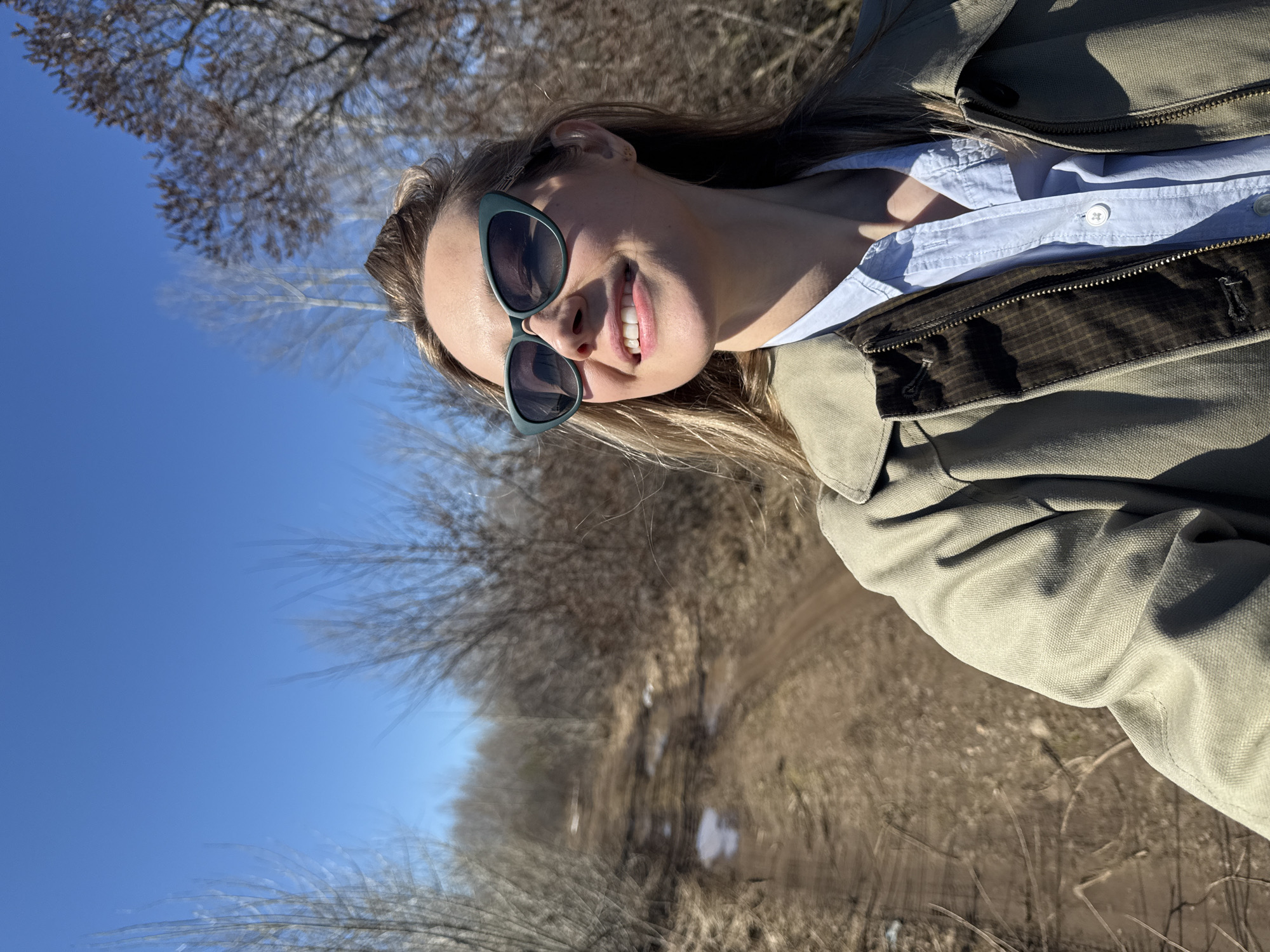 A smiling woman with sunglasses stands outdoors. She wears a light blue button-down shirt and a khaki-colored jacket. The background consists of a dirt path lined with bare trees, a patch of snow, and a clear blue sky. The location is Chaikovsky, Russia.