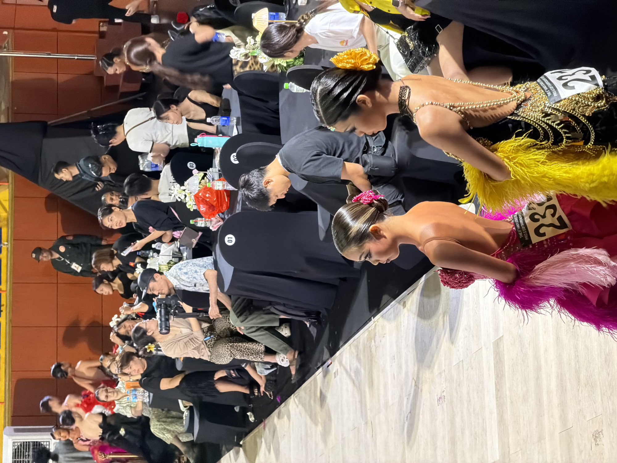 In a ballroom setting, likely part of a dance competition venue in Hanoi, Vietnam, individuals are seated and standing around tables draped with black tablecloths. Several people are observing a dance floor which is not fully visible. In the foreground, two young women in elaborate dance costumes, one in pink and one in black with intricate gold embellishments, are partially visible from the side and back, facing towards the dance floor. They are wearing numbers on their costumes, indicated by the visible "23" on both. The atmosphere appears to be one of anticipation or post-performance rest for the dancers. Spectators, including adults and possibly some younger individuals, are seated at tables, some holding phones and bottles of water. A man in a dark shirt is captured holding a camera, suggesting a media presence. In the background, a security guard in uniform stands near a red-orange wall, adding to the sense of an organized event. The lighting is typical of an indoor venue, with spotlights likely illuminating the dance floor area.