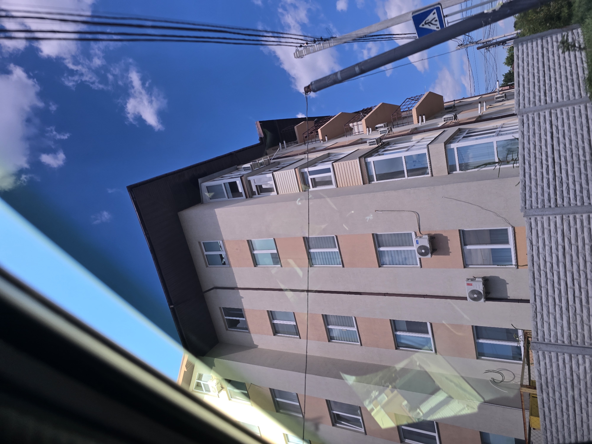 The media file depicts a view of a multi-story building from a low angle, possibly from a car, with reflections on a surface in the foreground. The building appears to be residential, with numerous windows, some of which have air conditioning units mounted outside. The facade is a mix of light beige and peach-colored panels, with dark brown trim along the roofline. The roof has several dormer windows and appears to be made of metal. The sky above is a clear, vibrant blue with scattered white clouds.
In the upper part of the image, power lines and a pole with a blue traffic sign featuring a pedestrian symbol are visible. To the right of the building, there is a grey, textured wall, possibly a fence or part of another structure, and some greenery suggesting trees. The overall scene implies an outdoor setting during the daytime, under a sunny sky. The reflections on the foreground surface, which is out of focus and tinted with blue and green hues, suggest it might be a car windshield or a glass window. There is no explicit activity or event taking place in the image; it is a static view of a building and its surroundings. No text is clearly legible within the image, apart from the symbol on the traffic sign. The location context is provided as Petropavlivska Borshchahivka, Ukraine.