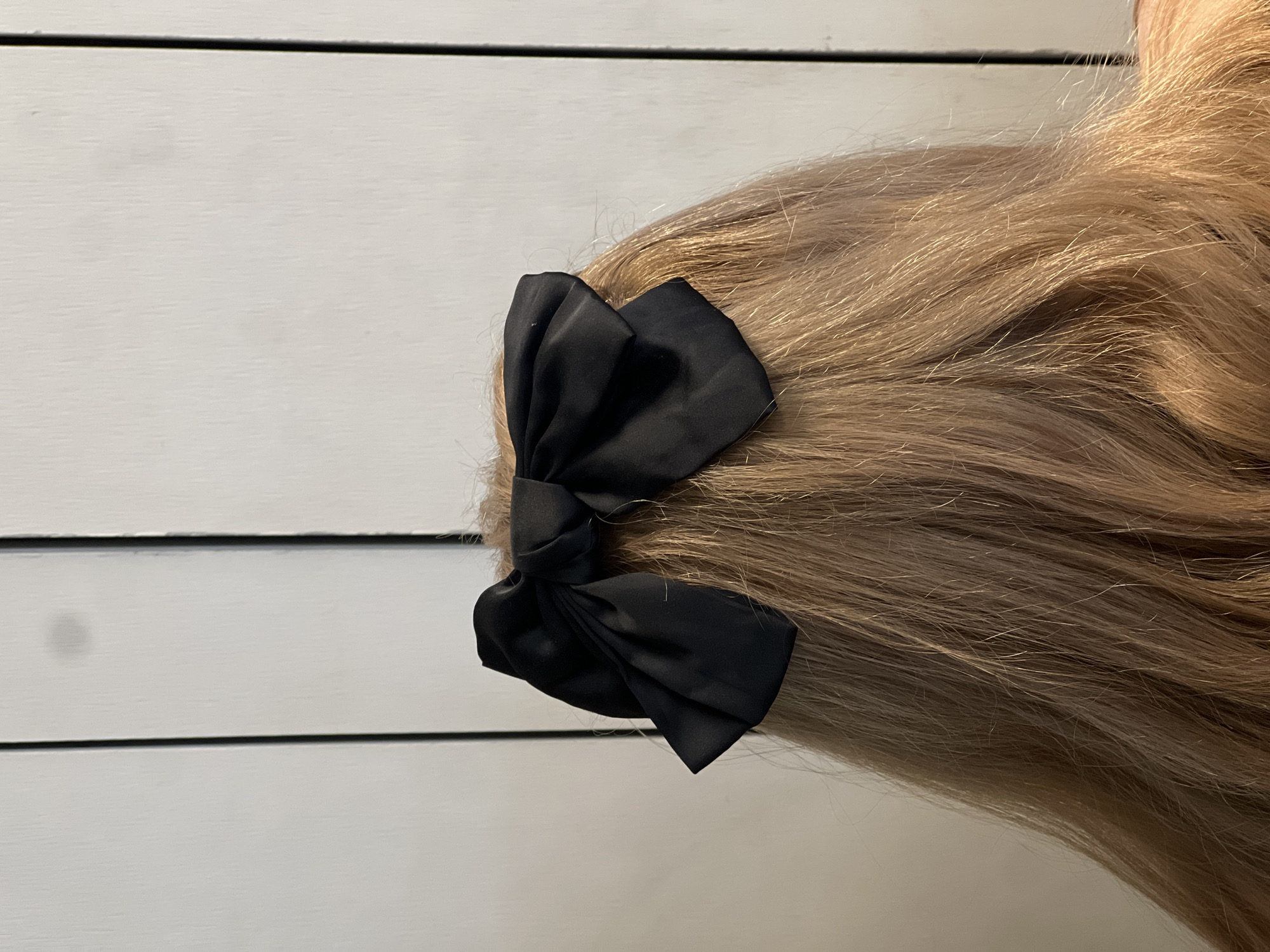 The image captures the back of an individual's head and upper shoulders, primarily focusing on light brown hair styled with a black fabric bow. The hair, appearing long and with subtle waves, displays variations in shade consistent with natural light brown or blonde tones. A black bow, constructed from what appears to be a soft fabric, is symmetrically tied and positioned centrally on the back of the head. The background consists of a light-colored, possibly white or off-white, surface featuring distinct vertical dark lines, suggesting paneling or seams, consistent with an interior wall or door. No other individuals, objects, or actions are discernible. The specific location within Tel-Aviv, Israel, cannot be determined from the visual information provided in the image.