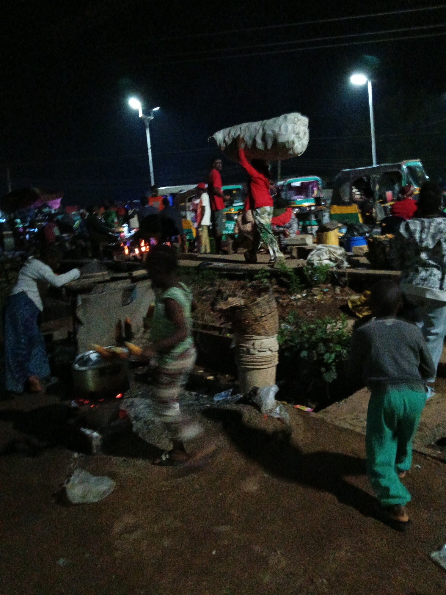 The image depicts an outdoor scene at night, likely in Bukuru, Nigeria, with artificial lighting from street lamps illuminating the area. Several people are visible, engaged in various activities. In the foreground, a person in a grey shirt and green pants is walking away from the camera, casting a long shadow. To the left, two individuals are near a cooking setup with a pot on a fire, where corn appears to be cooking. One person, wearing a white top and a blue skirt, leans over the cooking area, while a child in a green and white striped shirt stands nearby.
In the background, a person in a red shirt is carrying a large, white, cylindrical object on their head. Several auto-rickshaws, a common mode of transport in Nigeria, are parked or moving in the scene, some with colorful canopies. Other people are scattered throughout the scene, some appearing to be vendors or shoppers at what seems to be a market. The ground is a mixture of dirt and what looks like pavement. The overall impression is of a lively night market or street scene, possibly with food vendors and general commerce taking place. The lighting suggests it is nighttime, and there are no clear indicators of weather conditions. No visible text is present in the image.