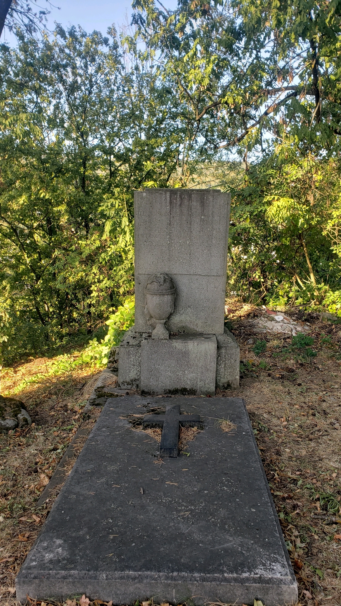 The image depicts a grave in a cemetery, likely located in Prešov, Slovakia, given the context. The primary subject is a tombstone constructed of grey stone. In the foreground, a large, flat stone slab forms the base of the grave, with a dark cross embedded on its surface. Behind this slab, a more elaborate monument rises, featuring a tall, rectangular headstone with faded inscriptions, flanked by smaller stone blocks. To the left of the headstone, a small, urn-like stone ornament is affixed. The surrounding environment is natural, with dry leaves and pine needles scattered on the ground. Dense foliage from trees with green leaves forms the backdrop, with a clear blue sky visible above. The lighting suggests it is daytime, with sunlight casting shadows and highlighting the textures of the stone and foliage. The scene is static, with no people or animals present.