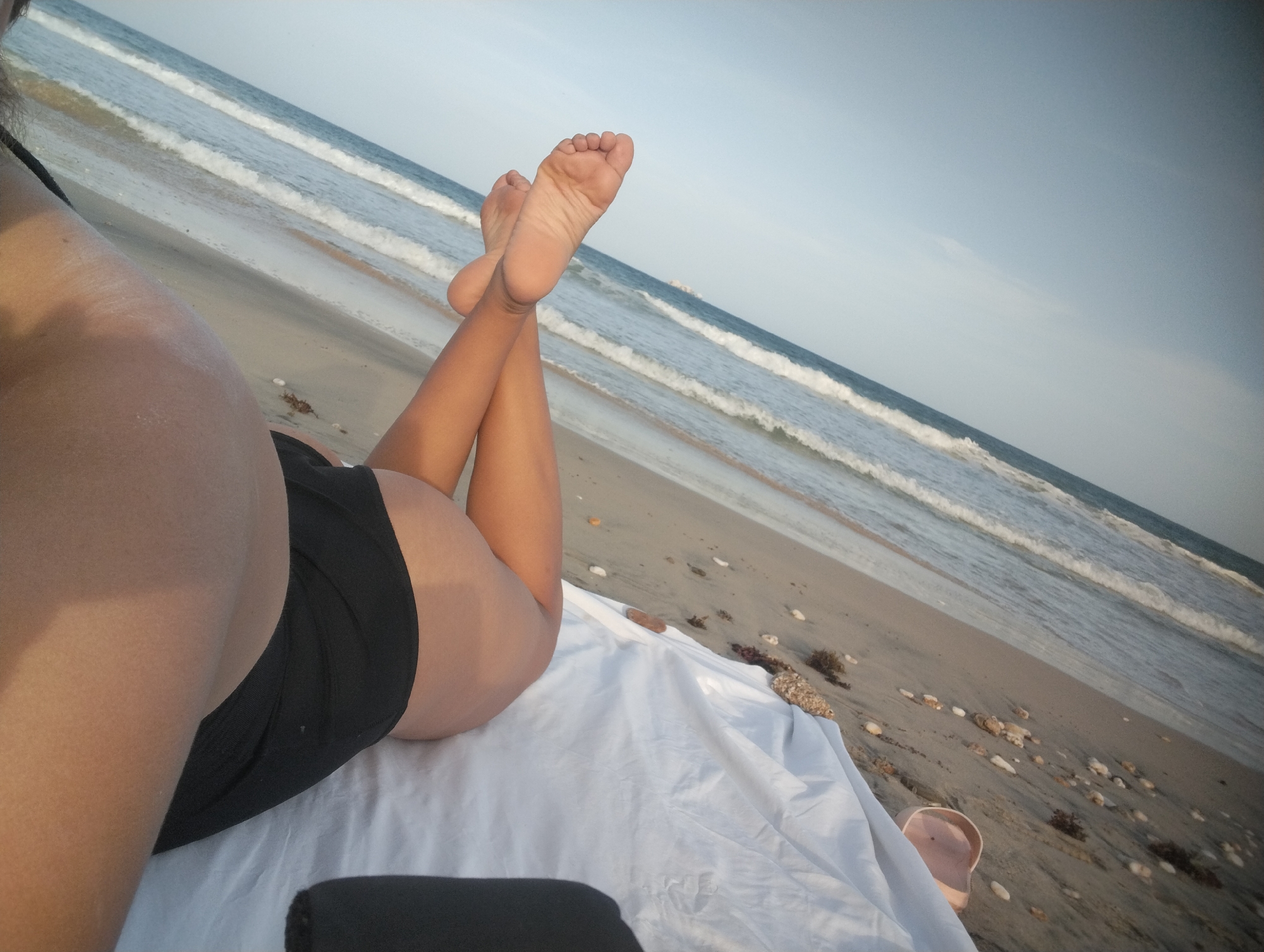 A person lies on a white sheet on a sandy beach. The person is wearing black swim bottoms. The person's bare feet are elevated with the ocean as a backdrop. Scattered seashells and debris are present on the sand near the sheet. The location appears to be a beach, possibly near Porlamar, Venezuela, considering the request.
