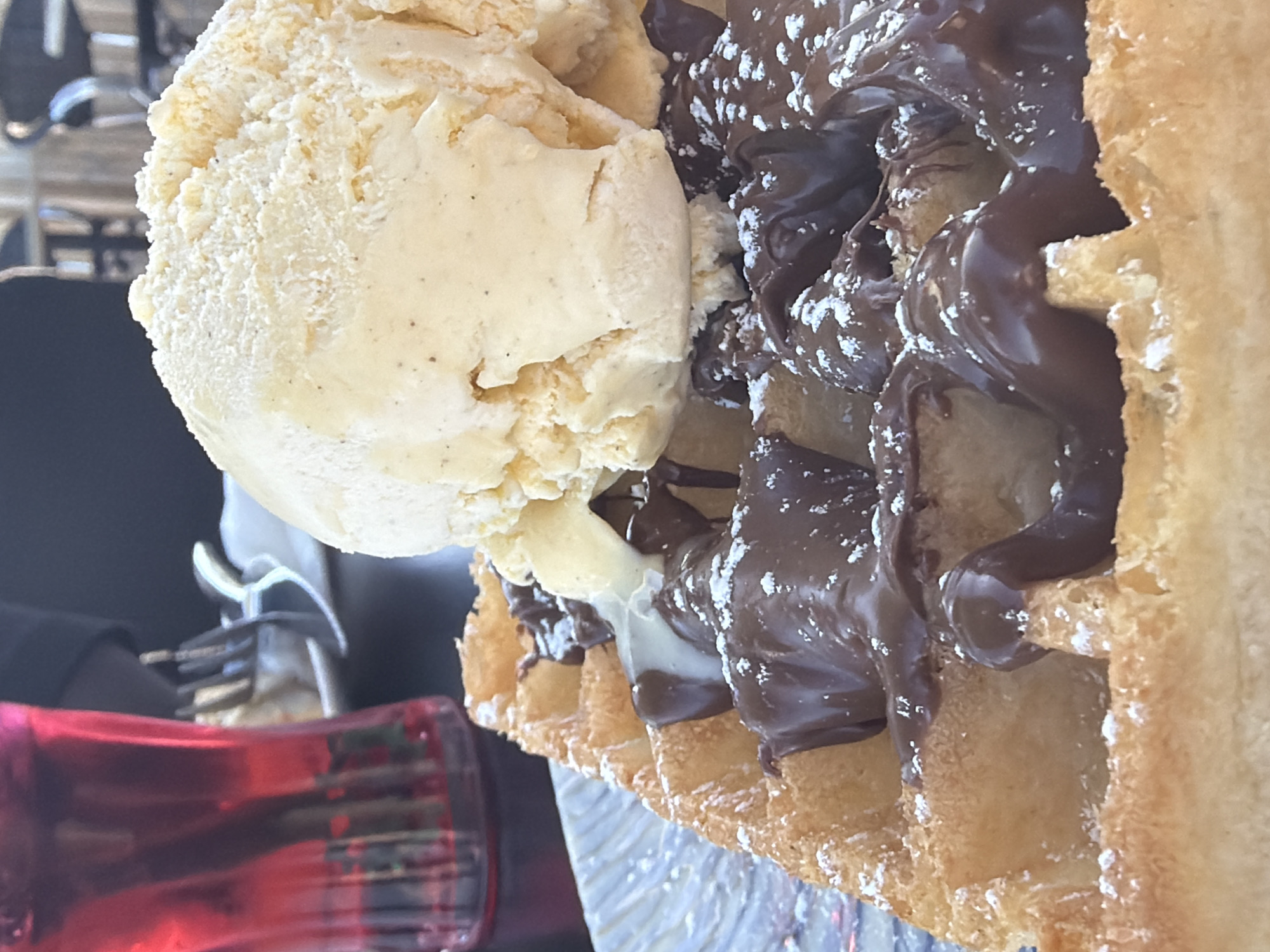 The image, captured in a close-up and rotated clockwise, features a tempting dessert: a waffle generously topped with a large scoop of ice cream and a thick layer of chocolate spread. The light yellow ice cream, possibly vanilla given the visible dark specks, is melting slightly, creating creamy drips down the side. A rich, dark brown chocolate spread, resembling Nutella, covers a significant portion of the golden-brown waffle, filling its grid indentations. Both the chocolate and sections of the waffle are dusted with white powdered sugar.
The waffle exhibits a crisp, inviting texture. In the blurred background, a translucent red object, possibly a drink container or glass, is visible towards the bottom left. Further back, out-of-focus metallic structures suggest an outdoor or cafe setting, potentially in Toulon, France, based on the provided context. The bright lighting indicates it is daytime. No people or legible text are visible in the frame.