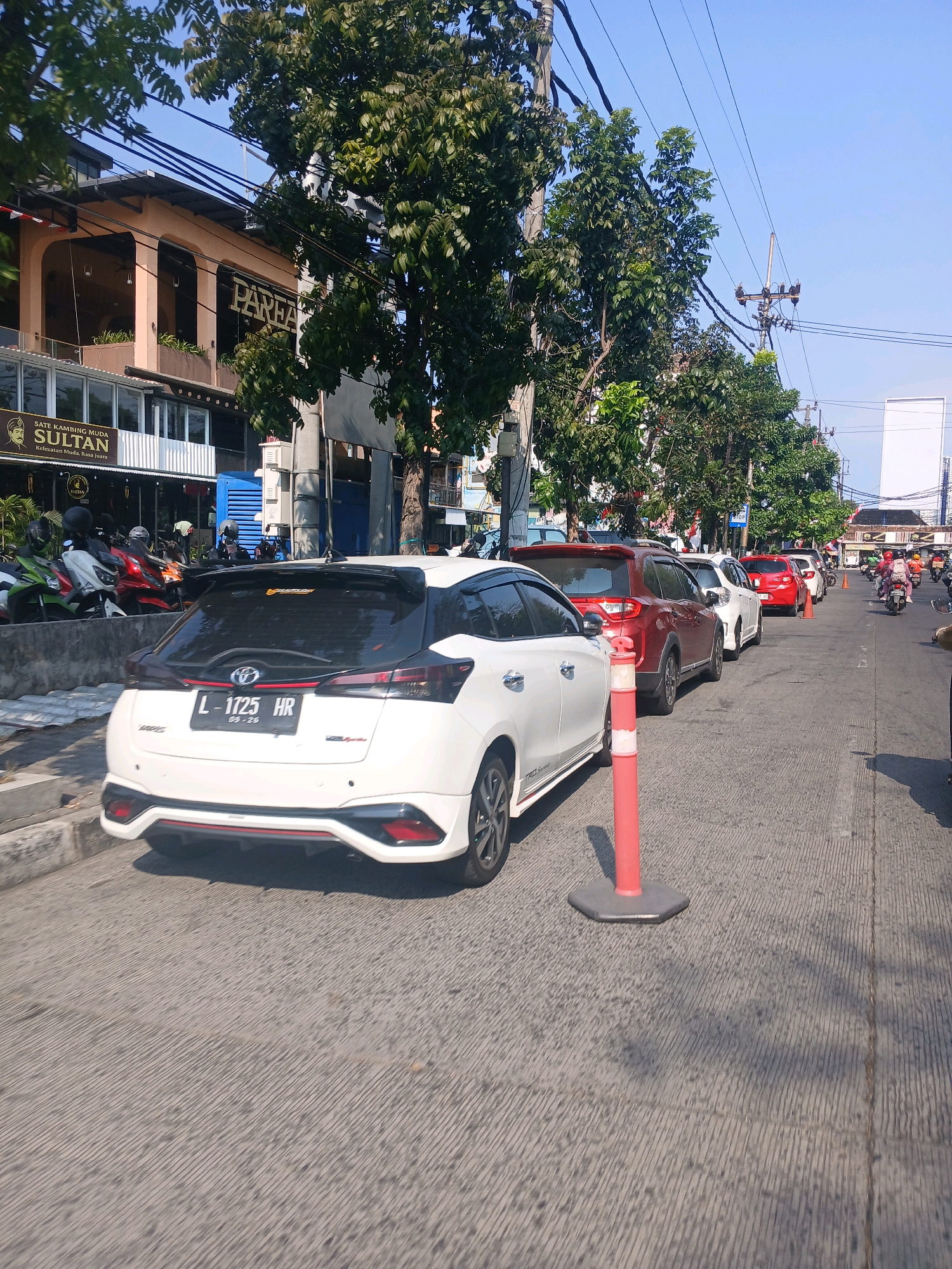 The image depicts a street scene in Surabaya, Indonesia, on a clear, sunny day. In the foreground, a white Toyota Yaris is parked, its license plate visible as "L 1725 HR". The car has a sporty spoiler and red accents. Behind it, a line of cars is seen stretching down the street, including a red SUV, a white sedan, and another red car. A bright orange traffic cone with a gray base is positioned next to the white Yaris.
To the left of the street, a sidewalk runs alongside buildings. One building has signage that reads "SULTAN" with a graphic of a person's face, and another sign above it says "PAREA". Several motorcycles are parked on the sidewalk, some with helmets on their seats. Trees with lush green foliage line the street and sidewalk, casting shadows on the road. Overhead, numerous power lines crisscross the sky, supported by utility poles. In the background, more buildings and a white rectangular structure are visible. Further down the street, several people on motorcycles are in motion, and traffic cones suggest a potential lane closure or construction area. The overall atmosphere is that of a typical urban environment with moderate traffic.