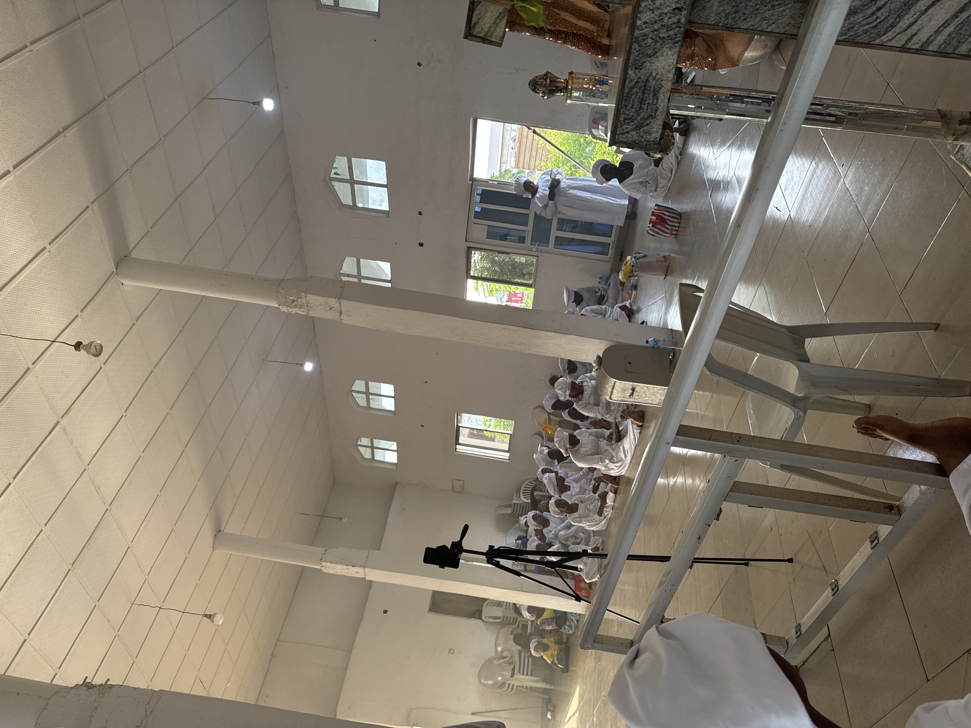 A group of people are gathered inside a building, possibly a church or community center, located in the city of Unknown, Unknown. The people are mostly dressed in white clothing and appear to be either praying or meditating, as many of them are kneeling or sitting on the floor. The building has white walls and a high ceiling with various windows that allow natural light to enter. There are some chairs arranged in rows where some of the people are seated. A video camera on a tripod is set up to record the event. A person's foot is visible in the foreground, suggesting someone is standing nearby.